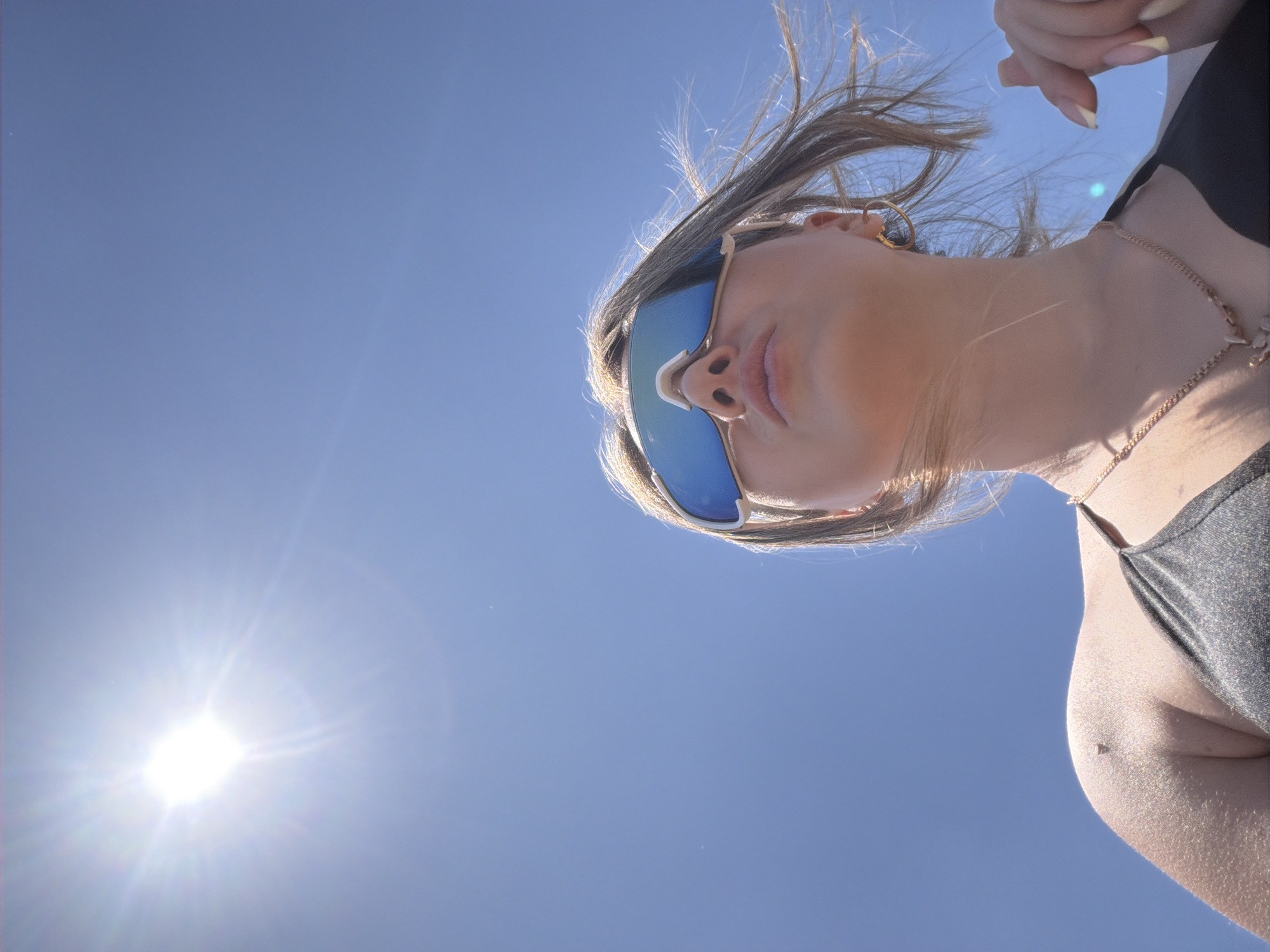 A low-angle, medium shot captures a woman from the chest up against a clear blue sky. The sun, positioned in the upper left quadrant, is intensely bright with visible rays. The woman is wearing large, blue-tinted wraparound sunglasses with white frames, obscuring her eyes. Her nose and lips are visible, and her blonde hair, windswept, is framing her face. She is wearing a dark, shimmering bikini top and a delicate gold necklace with a small pendant. Her left shoulder and upper arm are visible, showing sun-kissed skin and a small mole. Her right hand is partially visible in the lower right corner, with pale yellow painted fingernails. The overall environment is outdoors under a bright, sunny sky, suggesting a warm climate or season. There is no indication of the city of Shymkent, Kazakhstan.