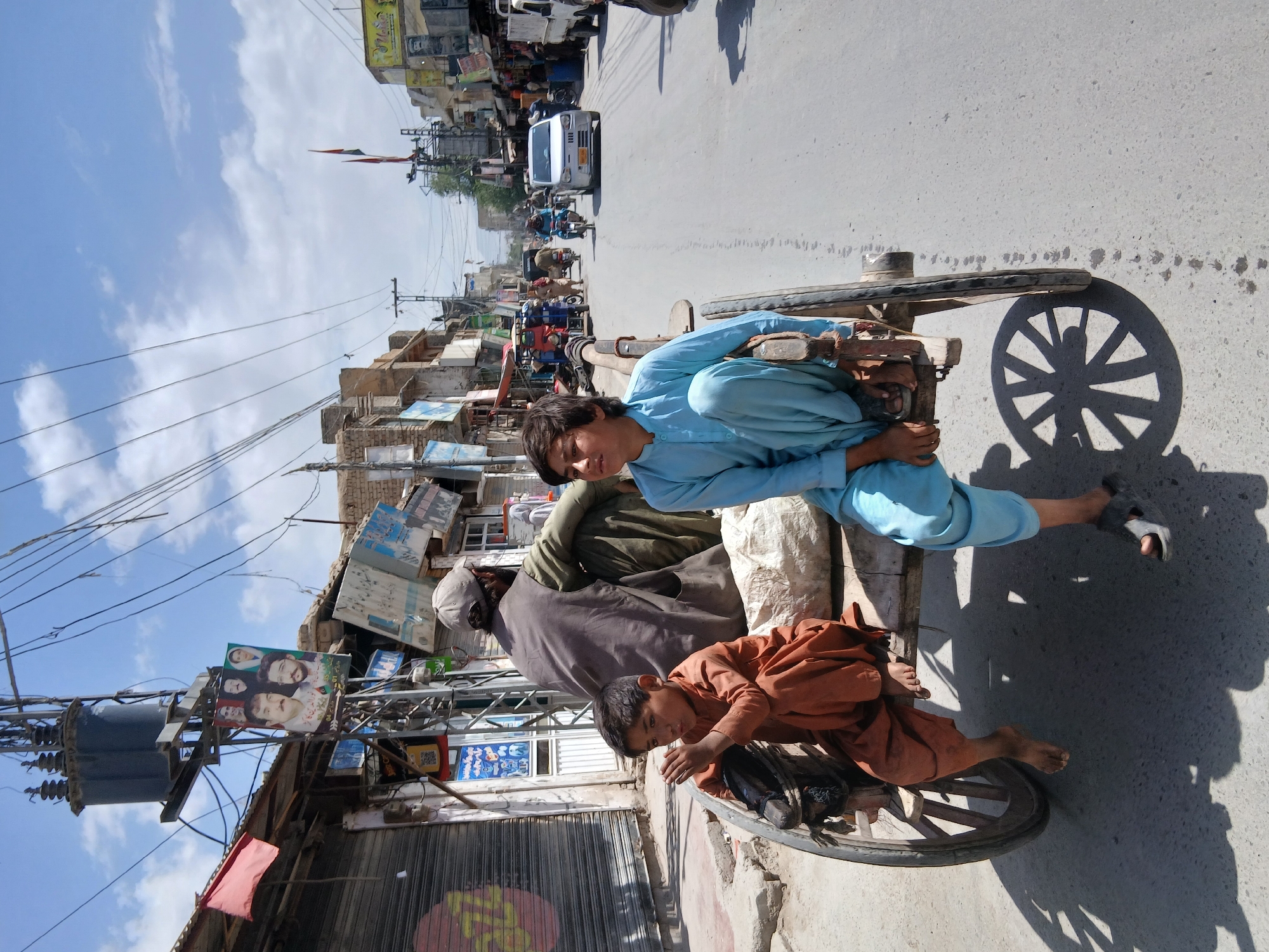 This image, rotated 90 degrees clockwise, captures a lively street scene in Quetta City Tehsil, Pakistan, under a bright, sunny sky with scattered clouds. The perspective is slightly elevated, looking down onto a bustling street.
In the foreground, three individuals are seated on and around a two-wheeled wooden cart. A young boy, wearing a light blue shalwar kameez, sits prominently on the cart, looking towards the camera with a calm expression. Below him, a younger boy, barefoot and dressed in an orange-brown shalwar kameez, sits near the cart's wheel, also observing the viewer. Behind the first boy, an adult male, partially obscured and wearing a grey outfit with a cap, is also seated on the cart.
The cart casts a prominent shadow of its spoked wheel on the light-colored asphalt road, indicating strong midday sunlight. The road extends into the background, where traffic flows. A white car, motorcycles, and other vehicles are visible.
Both sides of the street are lined with multi-story buildings, featuring shops at ground level with corrugated metal shutters and signage, and residential areas above. Utility poles heavily laden with a complex network of electrical wires crisscross the sky, with a large grey transformer mounted on one pole.
Various signs and banners adorn the buildings and poles. A notable large poster displays multiple faces, likely political figures, accompanied by text in an Urdu-like script, suggesting a political campaign. Another flag, likely the national flag of Pakistan, is also visible in the background. The scene depicts a slice of everyday urban life in a busy South Asian city.