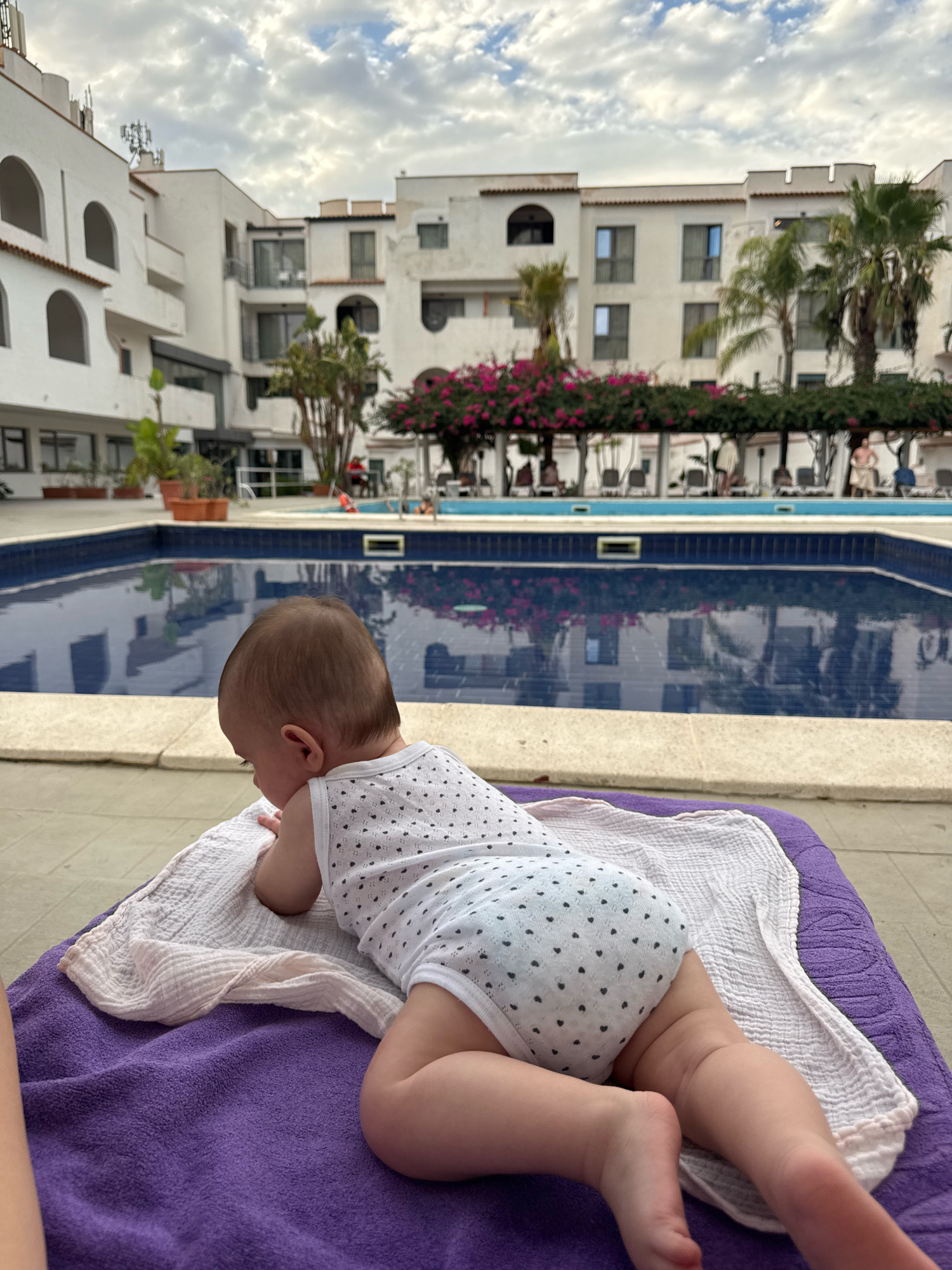 A baby is lying face down on a purple towel with a white cloth underneath it, positioned on a lounge chair. The baby is wearing a white onesie with small black hearts. Behind the baby is a swimming pool with a blue tiled bottom. The pool is surrounded by a light-colored stone border and a paved area. In the background, there are white buildings with arched windows and balconies, palm trees, and pink flowers. The sky is cloudy. The scene appears to be at a hotel or resort. The location is identified as being in the city of Isola delle Femmine, Italy.