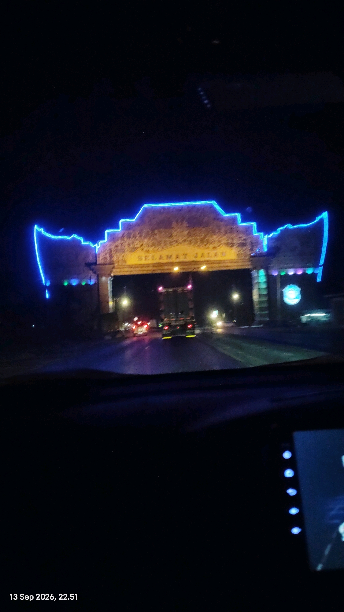 The image shows a nighttime view of an illuminated archway at the entrance to the city of Pandanan, Indonesia. The archway is decorated with bright blue neon lights that outline its structure, and it has text that reads "SELAMAT JALAN" in yellow lettering. Below the archway, a road is visible with traffic, including a large truck. The scene appears to be captured from inside a vehicle, indicated by the windshield's reflection and the edge of a dashboard on the right side of the frame. The overall ambiance is that of a well-lit city entrance at night.