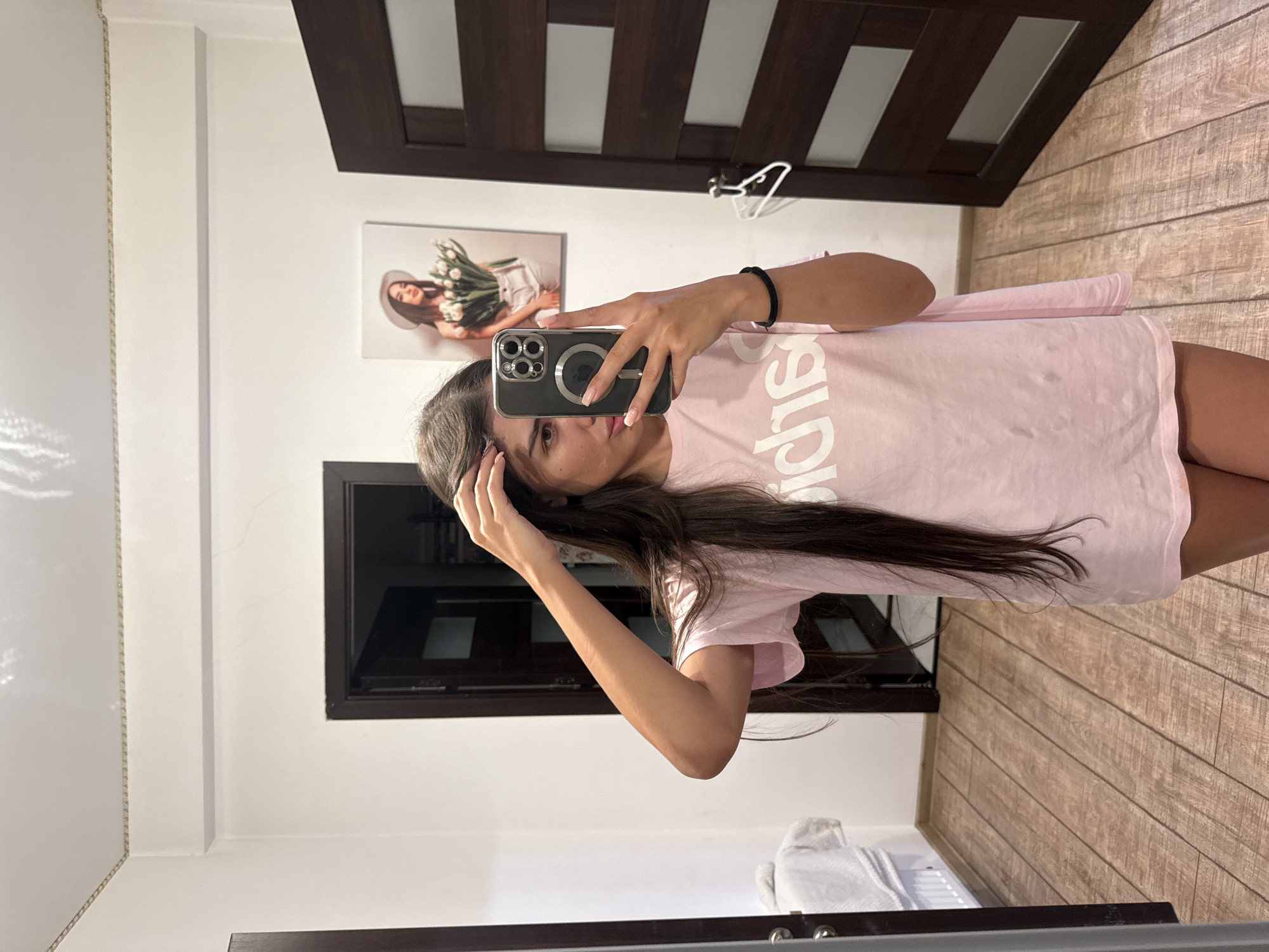 A young woman with long dark hair is taking a mirror selfie in a room. She is wearing a pink t-shirt with white lettering. She is standing in front of a dark wooden door, and to her left, there is a mirror. On the wall behind her, there is a framed picture of a woman wearing a hat and holding flowers. The room has wooden flooring and white walls. The environment suggests a residential setting, possibly a bedroom or a dressing room, in the city of Chișinău, Moldova.