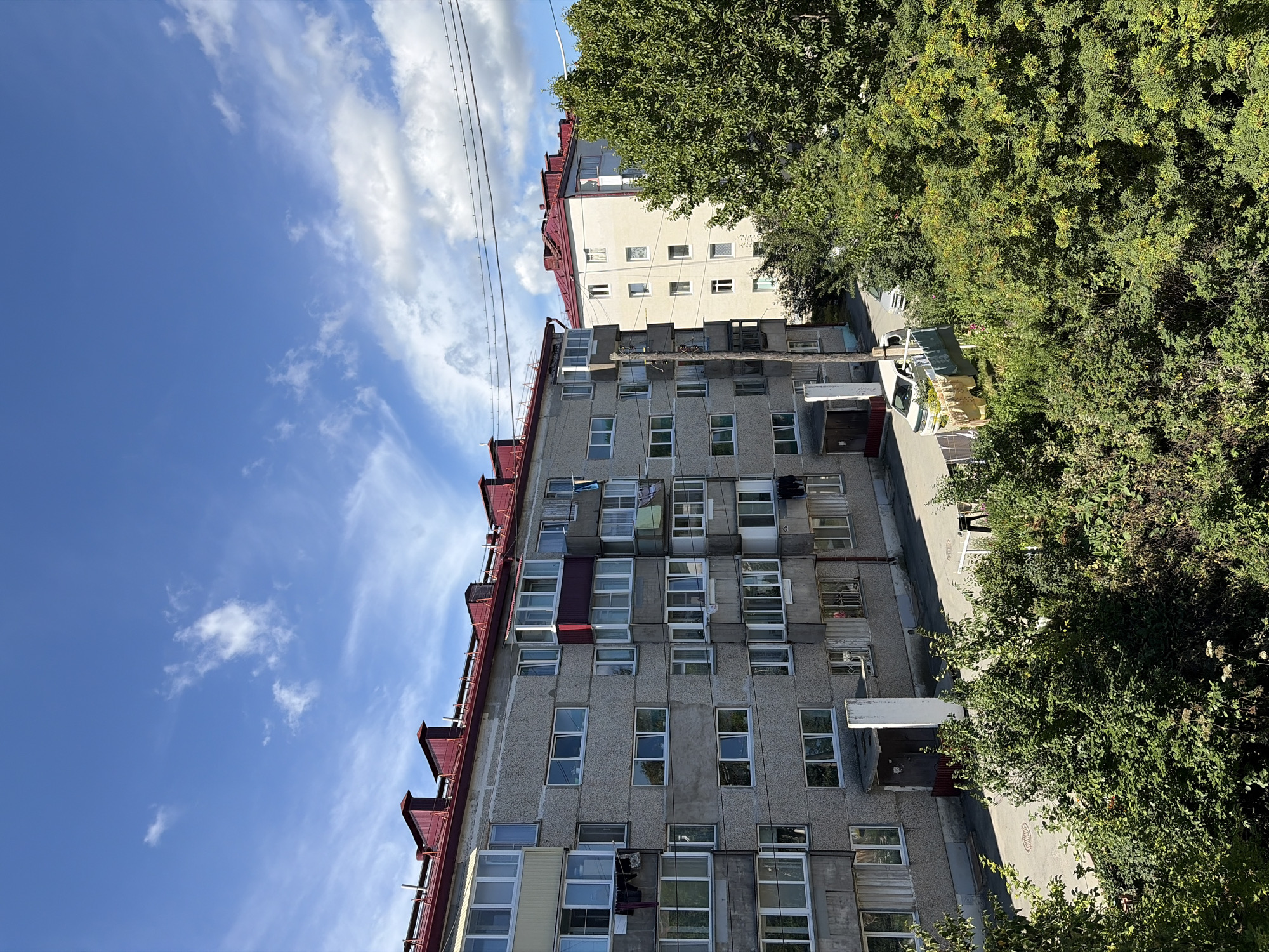 This image depicts a residential area in Korsakov, Russia, under a bright, partly cloudy sky. The foreground is dominated by lush green trees and bushes, obscuring the lower portions of two apartment buildings. The apartment buildings themselves are concrete structures with red metal roofs and numerous windows. Laundry is visible hanging from clotheslines on balconies of the left building, and a white car is parked in front of the buildings. The building on the right is lighter in color and appears to be of a similar architectural style. Power lines are strung across the upper portion of the image. The scene appears to be a typical urban residential setting on a clear day.