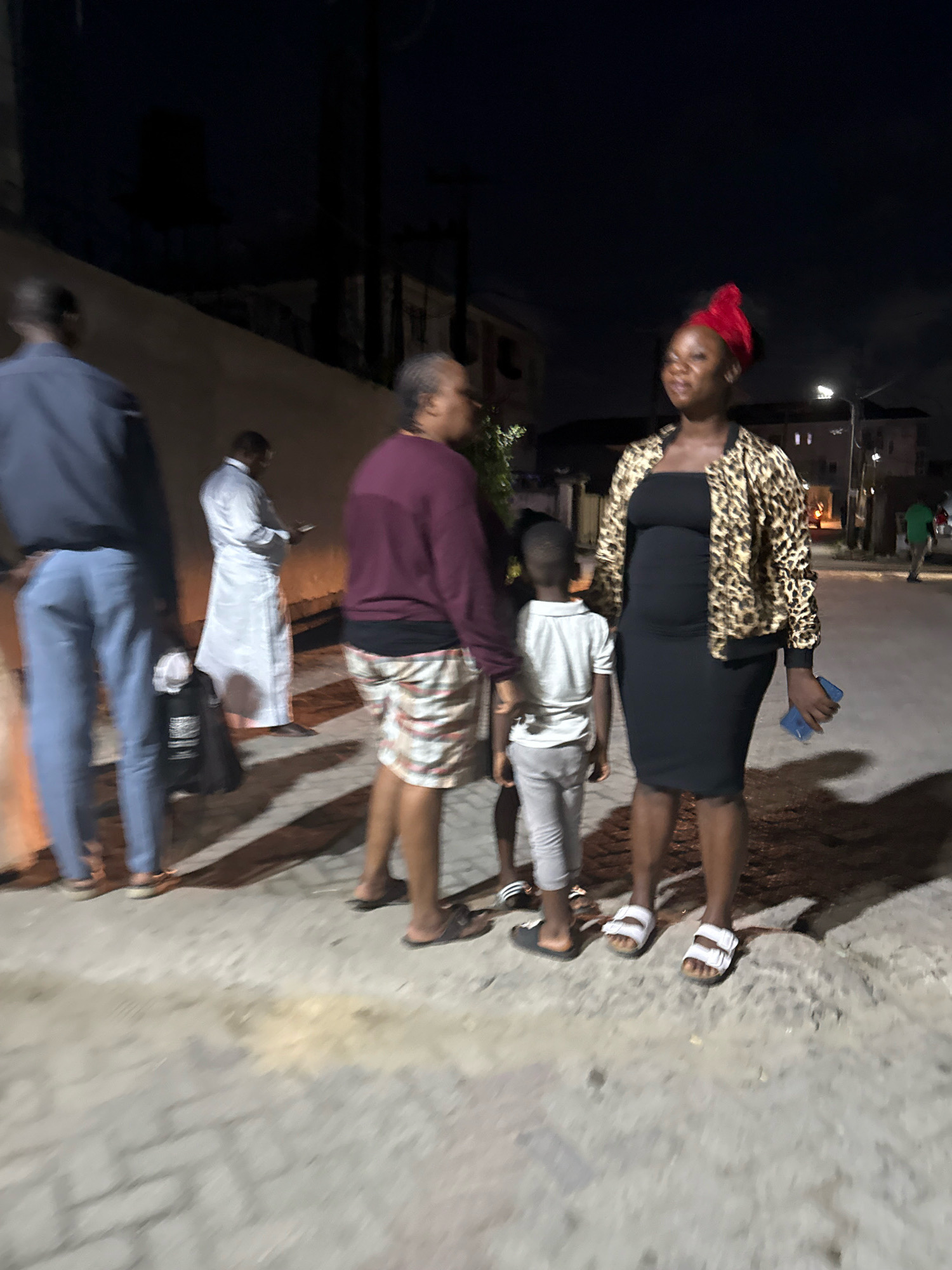 The image depicts a nighttime scene on a paved street in Ado, Nigeria. In the foreground, a woman wearing a black strapless dress, a leopard-print bomber jacket, and a red headscarf stands to the right. She holds a blue smartphone in her right hand and is wearing white sandals. Next to her on the left is a young boy in a white t-shirt and grey pants, also wearing sandals. To the left of the boy is another person, partially visible, wearing a maroon long-sleeved shirt and patterned shorts. Further to the left, a person in a dark blue shirt and light blue jeans stands with a black bag at their feet. In the background, a person in a long white garment stands and appears to be looking at a phone. The environment suggests an urban setting with buildings and streetlights visible in the distance. The lighting is dim, indicating it is night.