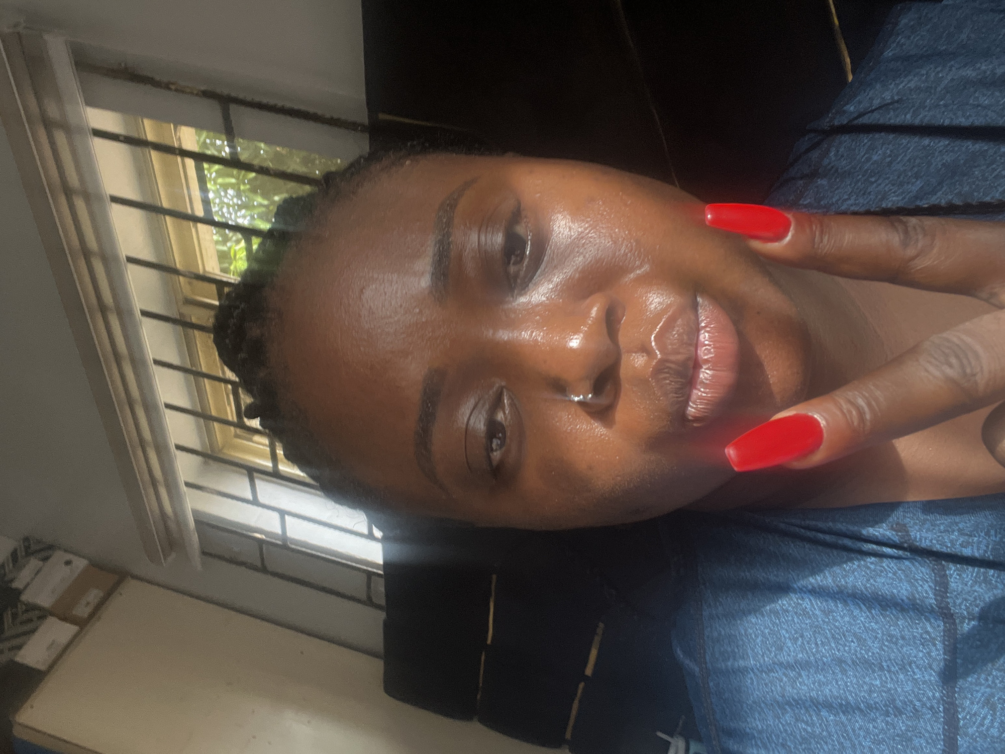 The image is a brightly lit, close-up selfie of a person, likely a young adult woman, with dark skin and dark hair styled in small braids. She is looking directly at the camera with a subtle smile or smirk, conveying a relaxed and positive mood. Her skin appears dewy or shiny, particularly across her forehead, nose, and lips, reflecting the strong light. Her eyebrows are well-defined.
She is wearing a blue garment, possibly made of a textured fabric. Her right hand is raised, with two fingers forming a 'V' or peace sign gesture. Both visible fingers have striking, bright red, square-tipped fingernails, adding a pop of color to the image.
The setting appears to be indoors, next to a window that allows ample natural light to stream in. The window features dark metal security bars, and through them, lush green foliage and bright outdoor sunlight are visible, indicating the presence of trees or plants outside. The strong, direct sunlight creates significant highlights on the subject's face and casts defined shadows. Portions of a light-colored wall or ceiling are visible at the top, and indistinct light-colored objects, possibly boxes or furniture, are partially seen in the bottom left background.
The intense natural light suggests it is daytime, likely morning or afternoon. The combination of security bars, outdoor greenery, and bright sunlight is consistent with an indoor setting in a tropical or sub-tropical climate, aligning with the provided location context of Kubwa, Nigeria. There is no visible text in the image.