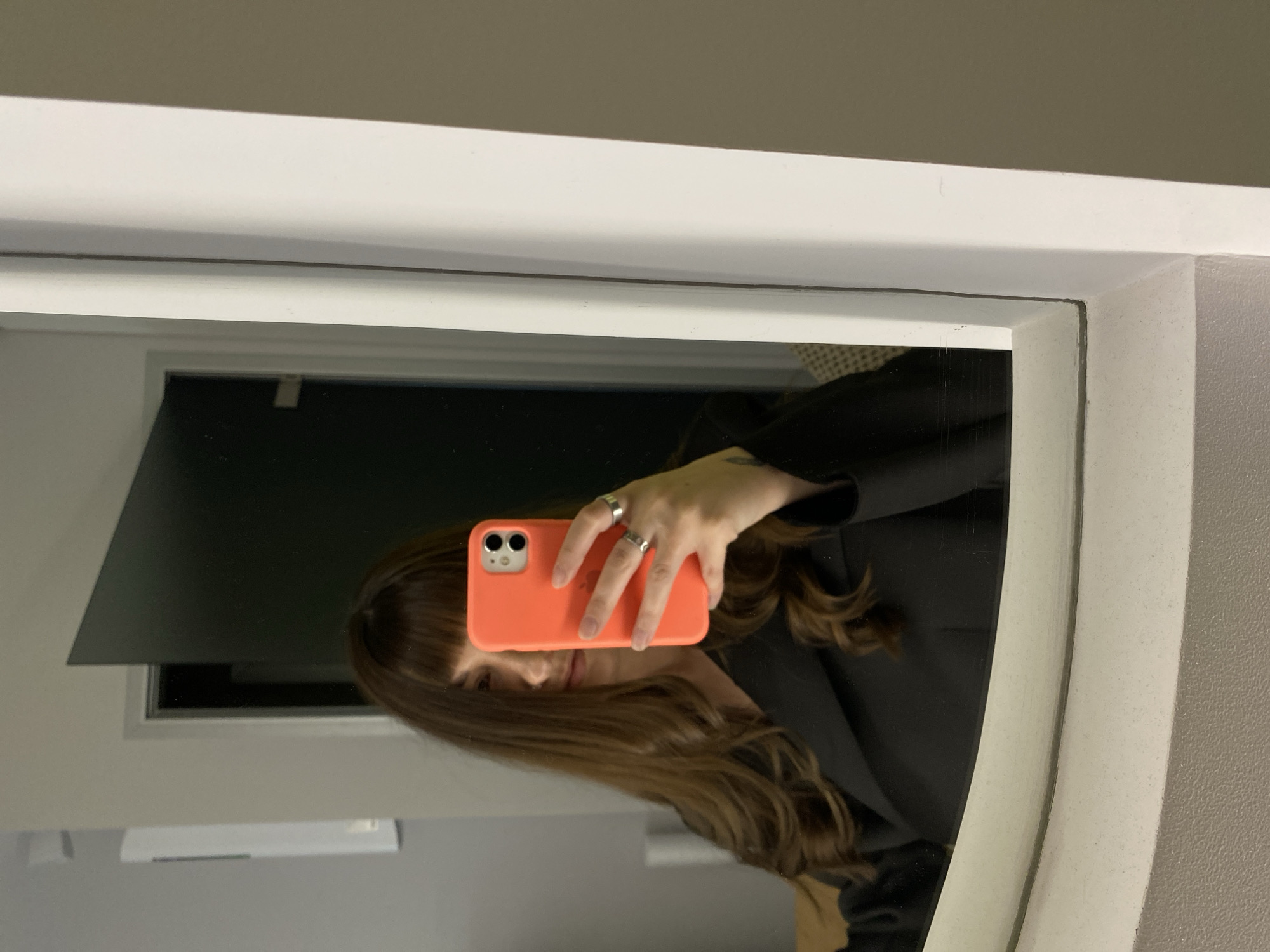 A person is taking a selfie in a mirror. The person has brown hair and is wearing a black blazer. The phone case is orange. A window is visible in the background. The location is unknown, but the user has specified Votkinsk, Russia.