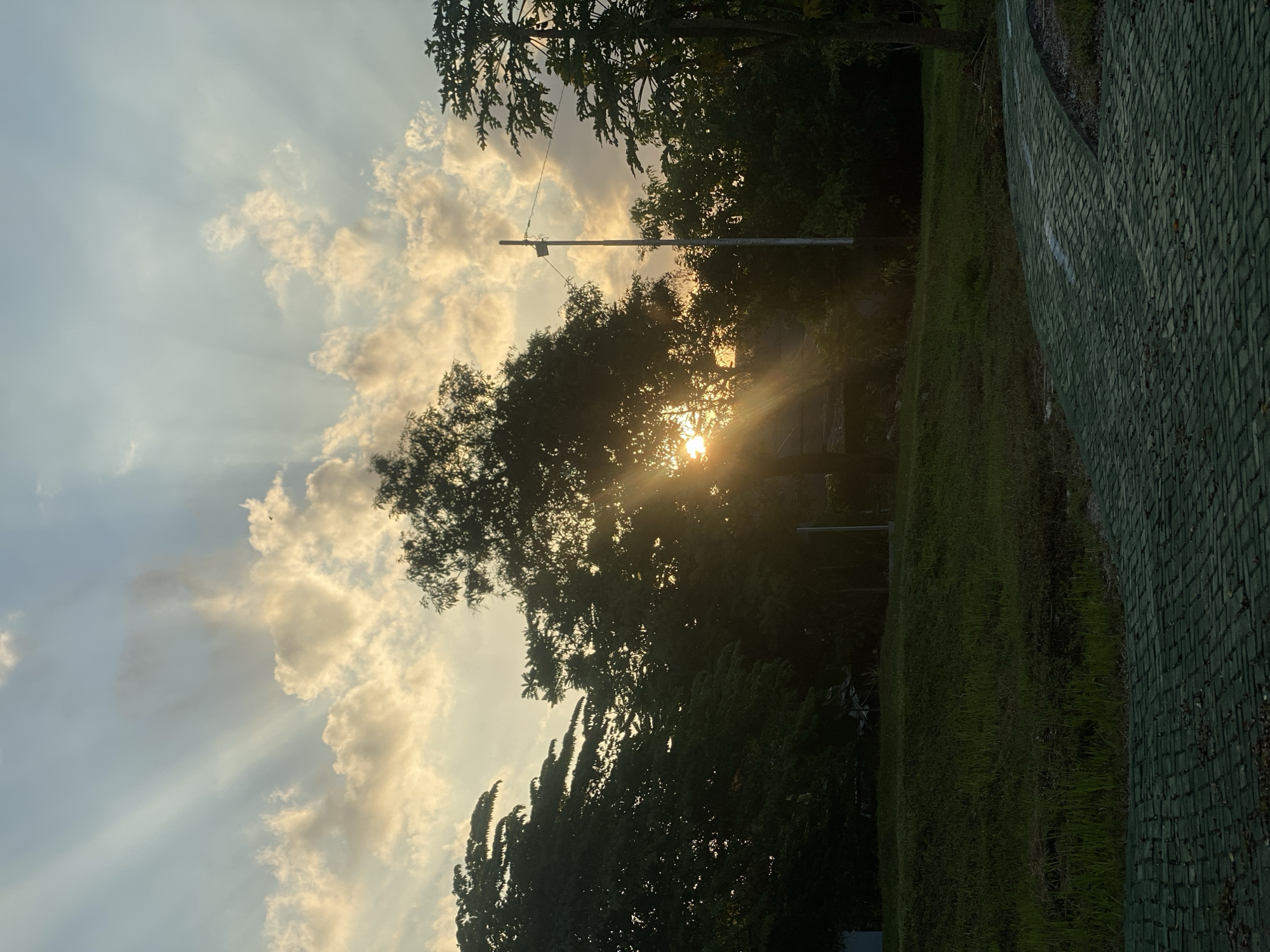 The image depicts a serene sunset scene in a park located in Banjarbaru, Indonesia. The sun is partially obscured by the silhouette of lush trees, casting dramatic sunbeams through the clouds. The sky is a gradient of soft blues and yellows, illuminated by the setting sun. In the foreground, a grassy slope leads to a paved pathway with a distinctive green, brick-like pattern. A utility pole stands on the right side of the frame, adding a touch of man-made structure to the natural landscape. The overall atmosphere is tranquil and picturesque, characteristic of a peaceful evening in a park.