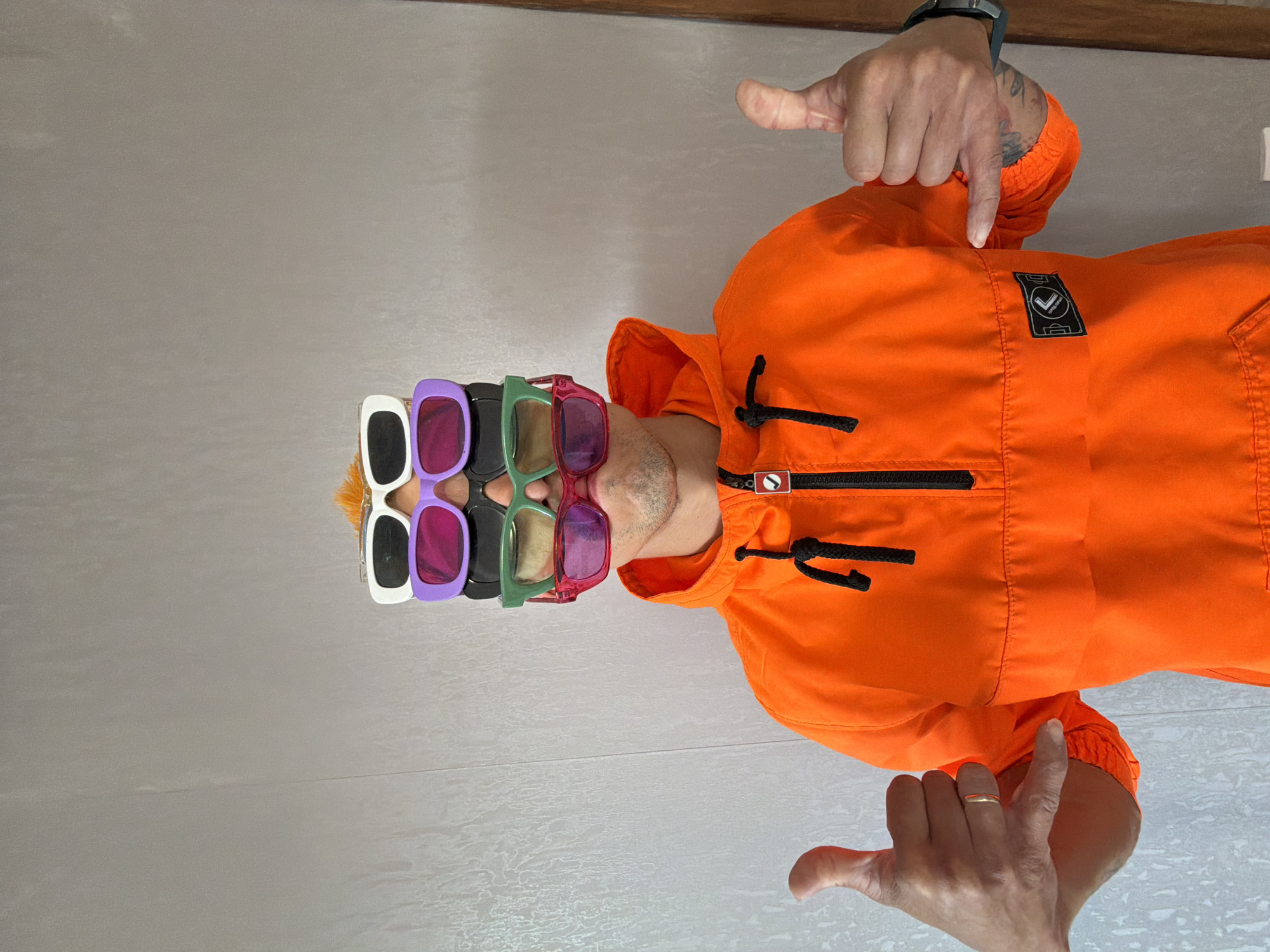 A person wearing an orange hooded jacket stands against a textured grey wall. They have four pairs of sunglasses stacked on their head, with the top pair being white, followed by purple, green, and pink. Their hair is an orangey-blonde mohawk visible above the sunglasses. Both hands are raised, with the left hand giving a thumbs-up and the right hand making a shaka sign. The person has visible tattoos on their right arm. The image is taken from the chest up. There is no visible indication of the location being in Chaikovsky, Russia.