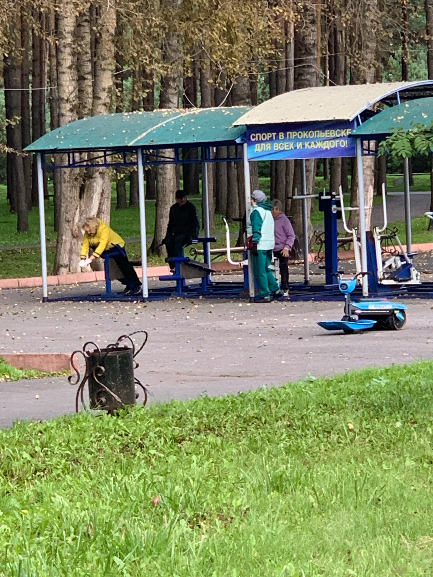 A man in a yellow jacket is exercising on a blue and metal workout machine under a covered outdoor gym area in Prokopyevsk, Russia. The gym equipment is set up on a gravel surface, surrounded by trees. Two women are standing nearby, one in a green tracksuit and the other in a purple jacket. A blue electric scooter is parked on the gravel in the foreground, next to a decorative metal trash bin. The gym has a blue banner with white text that reads "СПОРТ В ПРОКОПЬЕВСКЕ, ДЛЯ ВСЕХ И КАЖДОГО!", which translates to "SPORT IN PROKOPYEVSK, FOR ALL AND EVERYONE!". The area appears to be a public park or recreational space.