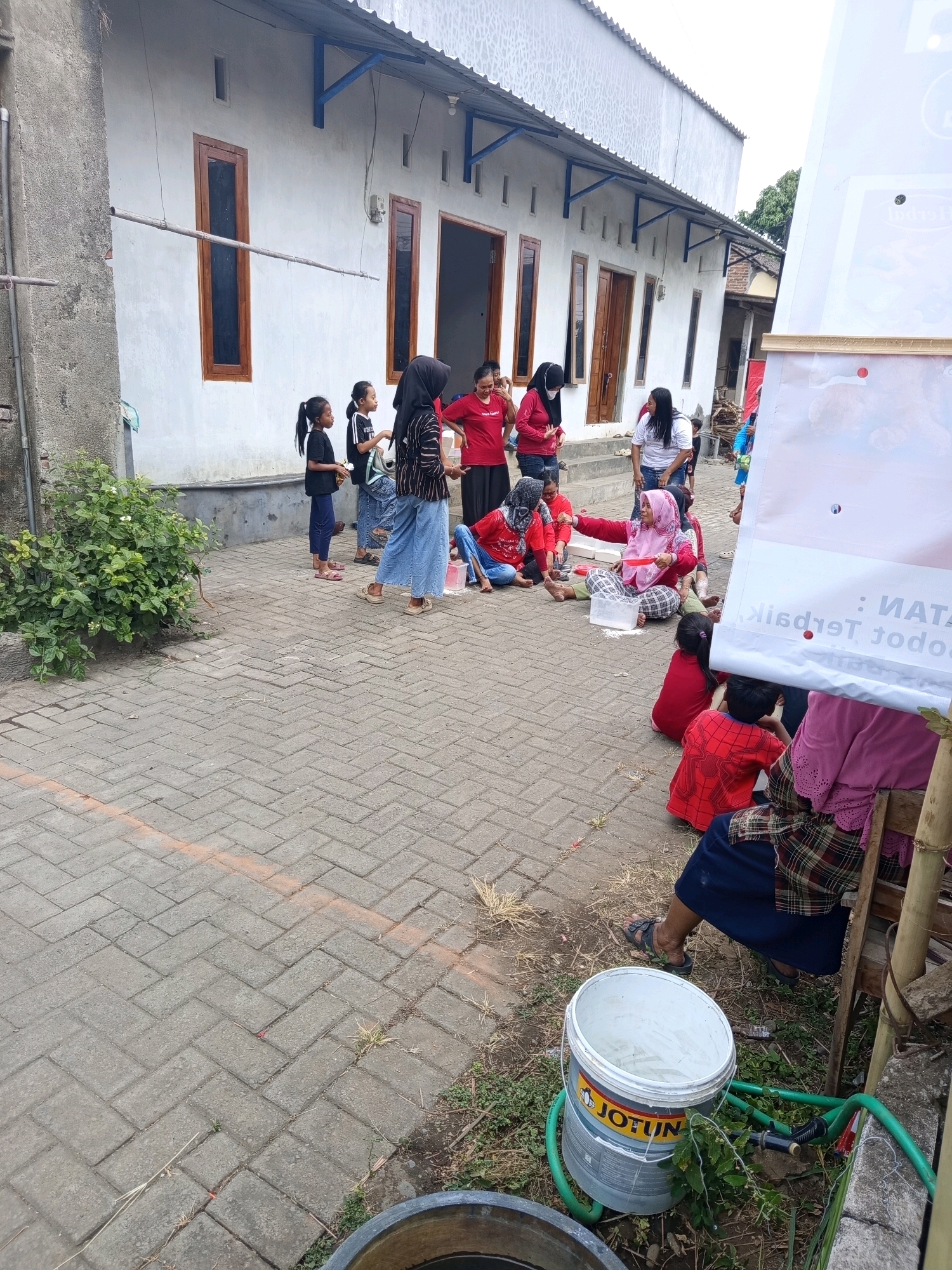 The image depicts an outdoor scene in the city of Ngablak, Indonesia, likely a residential area or community space, with a long, single-story building with white walls and wooden-framed windows. A group of people, including children and adults, are gathered on a paved brick walkway in front of the building. Some individuals are sitting on the ground, while others are standing. Several people are wearing red clothing, and some children are dressed in black tops and blue pants. A large white bucket with a green hose attached is prominently placed on the ground near the front right of the image. A partially visible banner or sign with text in Indonesian is also present on the right side. Green foliage and plants are visible on the left side and surrounding the scene, suggesting a somewhat unkempt or natural environment. The overall impression is of a casual gathering or activity taking place outdoors.