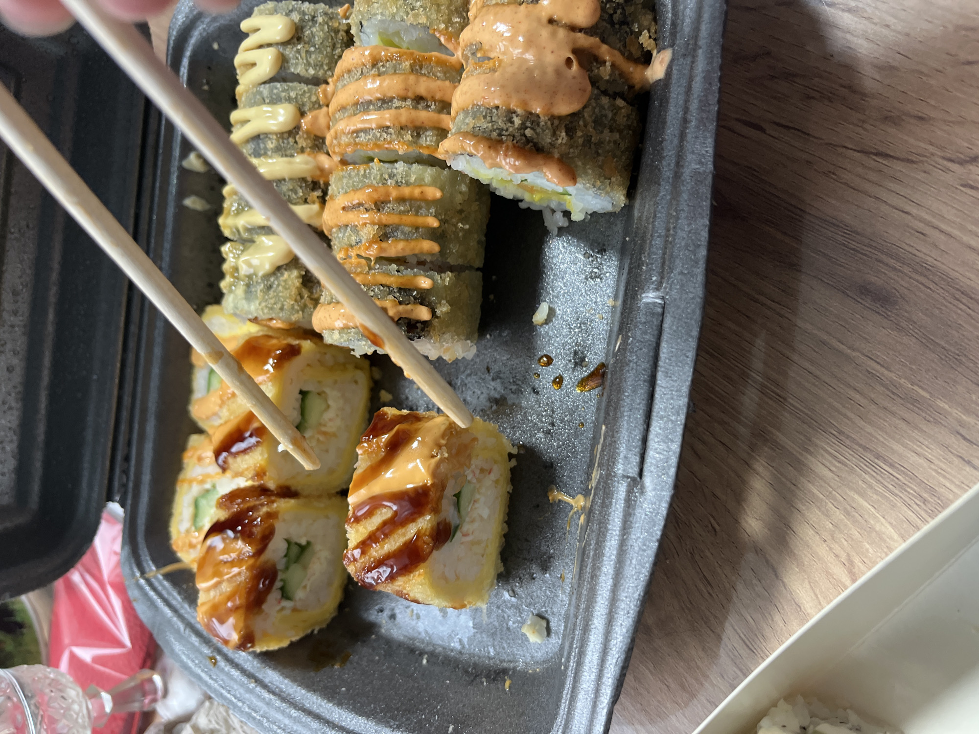 The image shows a close-up view of several pieces of sushi in a black takeout container. The sushi appears to be various types of rolls, some deep-fried and topped with sauces. Chopsticks are visible, one pair in the process of picking up a piece of sushi. The container is placed on a wooden surface, likely a table. In the background, a portion of a white container with rice is visible. There are no people clearly depicted, but the presence of chopsticks suggests someone is about to eat the sushi. The lighting is bright, indicating it is daytime, and the setting appears to be indoors. There is no visible text in the image. The overall impression is a casual meal being prepared to be eaten.