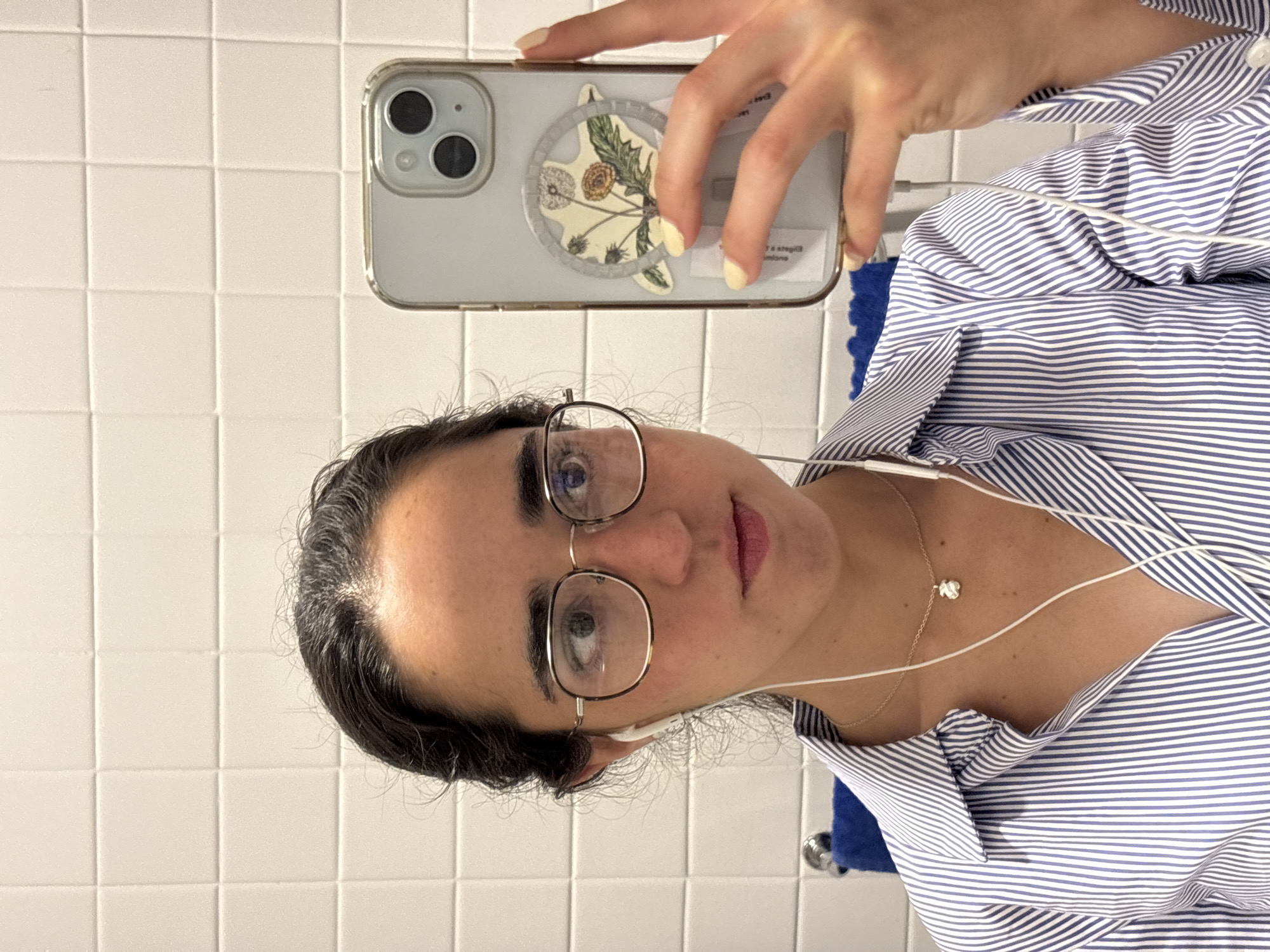 A woman takes a selfie in front of a white tiled wall. She is wearing glasses, in-ear headphones, and a blue and white striped shirt with a delicate necklace featuring a small white flower. Her phone, which has a light blue case, is decorated with a flower sticker. The woman is positioned indoors, seemingly in a bathroom or similar space with white tiled walls. The location is within Mexico City, Mexico.