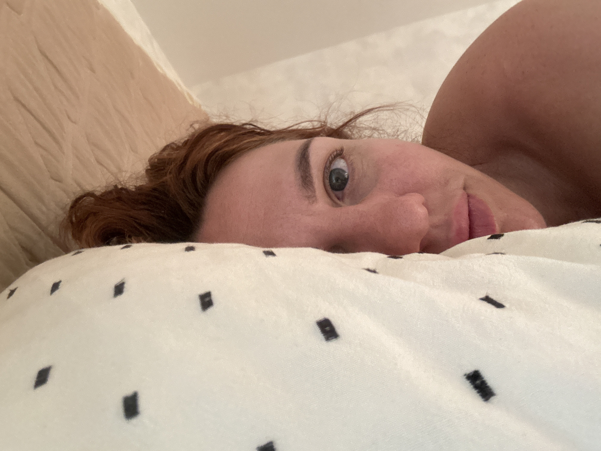 A close-up, high-angle photograph captures a person lying in bed indoors, likely in Sochi, Russia. The individual, appearing to be female, is shown from the perspective of someone looking down at them. Her reddish-brown hair is slightly disheveled, suggesting she may be waking up or resting comfortably.
The person's head rests on a white duvet or pillow cover adorned with a scattered pattern of small, irregular black squares. Her right eye is open and looks directly at the viewer, revealing a light-colored iris. The visible part of her face, including her forehead and the side of her mouth, shows a soft, gentle expression, possibly a hint of a sleepy smile. Her light-toned skin is visible on her face and shoulder, which appears in the upper right portion of the frame. Behind her head, a lighter, textured background, possibly a pillow or headboard, is visible. The scene evokes a tranquil and intimate moment in a bedroom setting. No text is visible in the image.