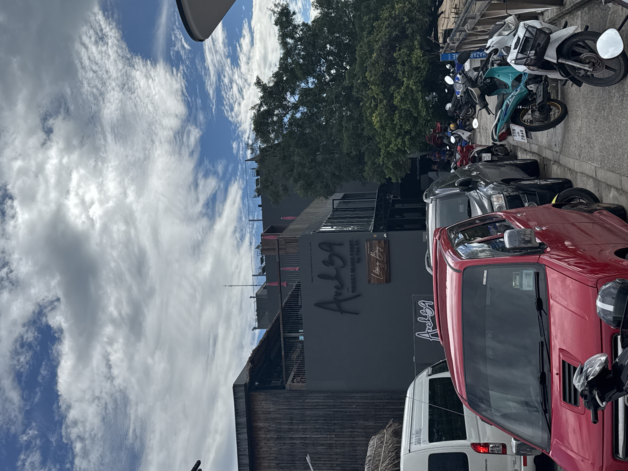 A low-angle outdoor shot captures a street scene in Chalong, Thailand, featuring a building with a prominent "Arch39" sign and parked vehicles. The sky is filled with dramatic clouds against a blue backdrop.
The building, a dark grey structure, has a modern architectural style with vertical wooden slats on the left side and what appears to be a thatched roof section. The "Arch39" logo is displayed in a stylized script on the building's facade, with smaller text beneath it reading "PHUKET BEACH FRONT By The EI" and a wooden sign with "Long Bay Cafe Phuket". Pink umbrella-like structures are visible on an upper level.
Parked vehicles line the sidewalk. In the foreground, a bright red car is visible, followed by a dark SUV. Beyond these, a variety of motorcycles are parked, including a prominent teal and white scooter. Some of the motorcycles have license plates. The ground appears to be a paved walkway.
The environment suggests a commercial area, possibly near a beach given the "Beach Front" signage. The lighting indicates it's daytime, with diffused sunlight due to the cloud cover. No people are clearly identifiable in the scene, although there are suggestions of individuals in the distance near the parked motorcycles.