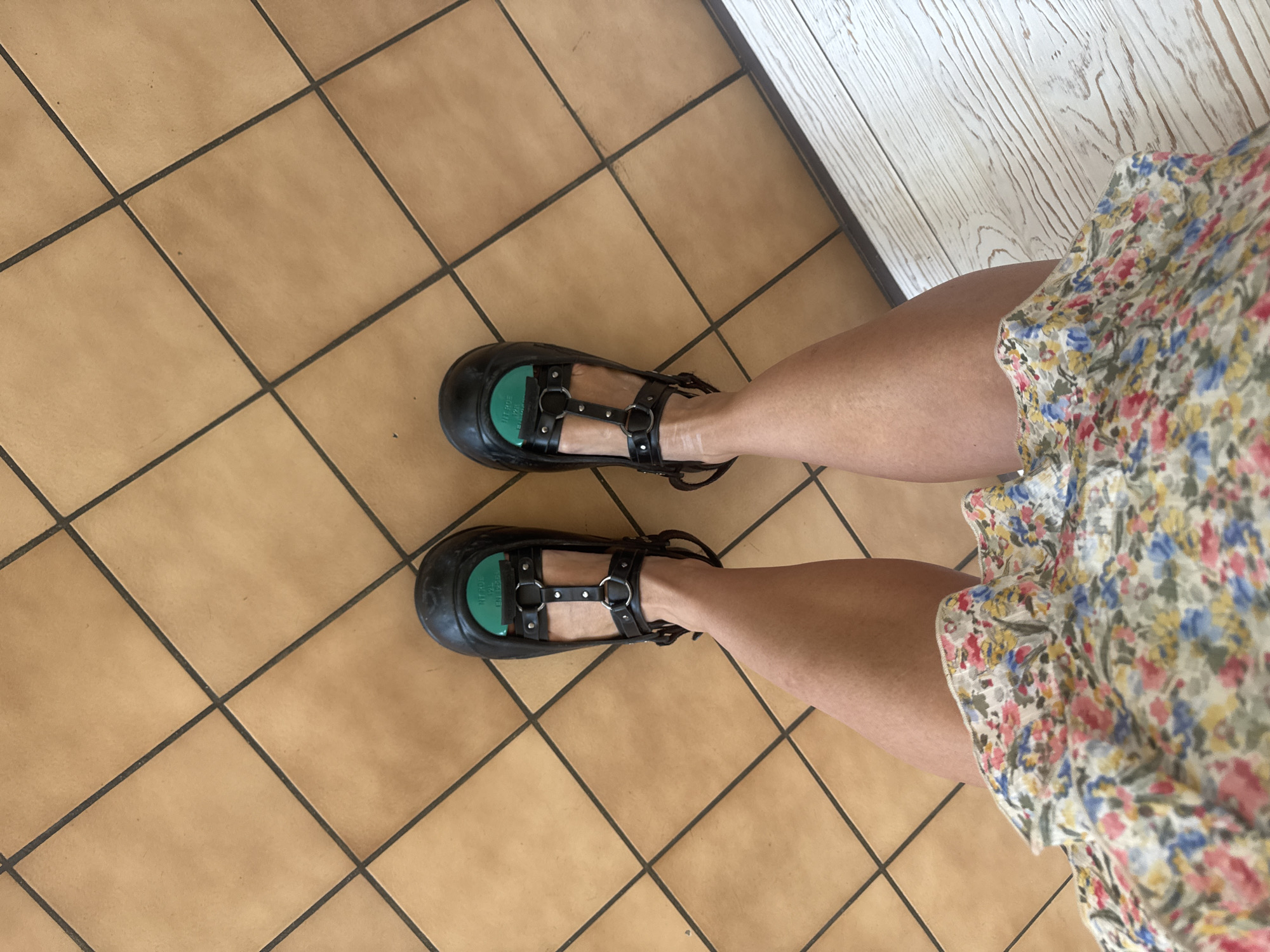 The image shows a person's legs and feet, viewed from above. The person is wearing black platform Mary Jane shoes with multiple straps and silver buckles. A green insert is visible inside each shoe. The person is also wearing a short, floral-patterned dress. The floor is tiled with large, square, light brown tiles, and there is a section of light-colored wooden planks to the right of the tiles. The image does not contain any information that would allow for the determination of a specific location in the city of Nivigne et Suran, France.
