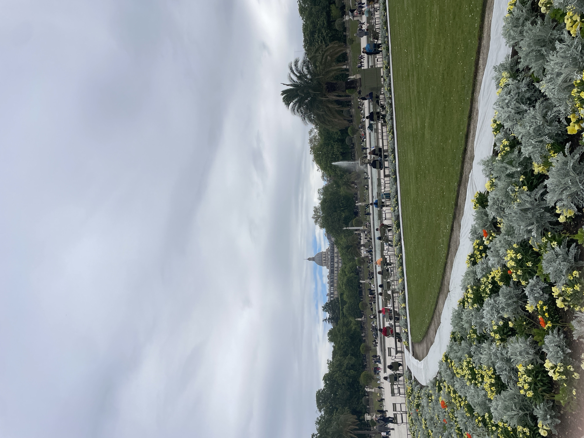 The image shows a view from the Jardin du Luxembourg in Paris, France. A symmetrical layout features a long green lawn bordered by rows of trees and chairs where people are sitting. In the distance is a building with a dome, potentially the Panthéon. A white gravel path runs along the edge of the lawn, lined with flowerbeds of silver foliage and yellow flowers, some with orange accents. The sky is overcast with gray and white clouds. A fountain is visible in the mid-ground, adding to the park's aesthetic. The scene reflects a relaxed atmosphere in an urban setting.