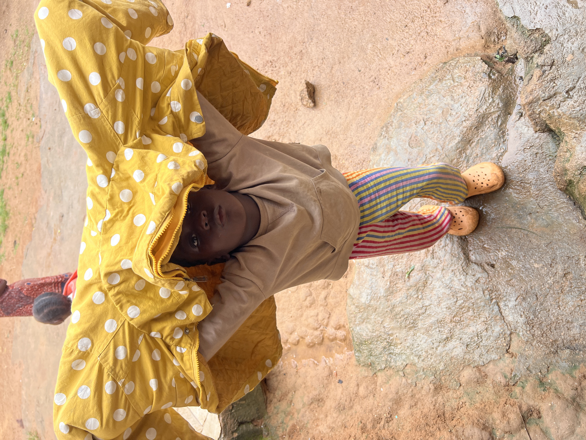 A young child, possibly female, stands on wet, uneven ground in what appears to be an outdoor, informal urban setting in Jos, Nigeria. The child is wearing a light brown long-sleeved shirt with a hood and colorful striped pants. They are holding a large, bright yellow jacket with white polka dots over their head, creating a makeshift shelter. The jacket is zipped up, and the child's face is visible peeking out from beneath it, looking directly at the camera with a neutral expression. In the background, out of focus, another person is partially visible, wearing a patterned red and black garment. The ground is a mix of reddish-brown earth and wet, greyish rocks. The overall impression is one of a candid moment captured in a humble environment.