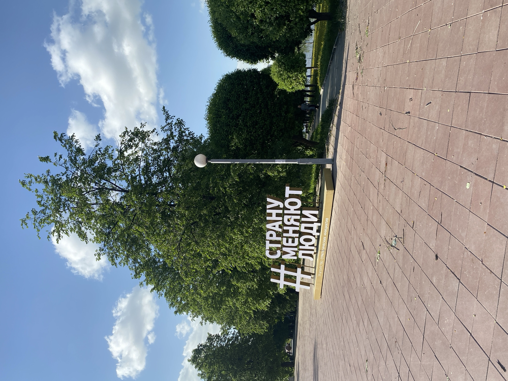 The image depicts a sunny day in Yekaterinburg, Russia. The ground is covered in red brick pavers, and a large metal sign stands on a short pedestal. The sign reads "#СТРАНУ МЕНЯЮТ ЛЮДИ" in white letters, which translates to "#PEOPLE ARE CHANGING THE COUNTRY". Behind the sign is a lamp post with a round white lamp and lush green trees with round foliage. In the background, other people can be seen sitting on benches along the path. Fluffy white clouds dot the blue sky.