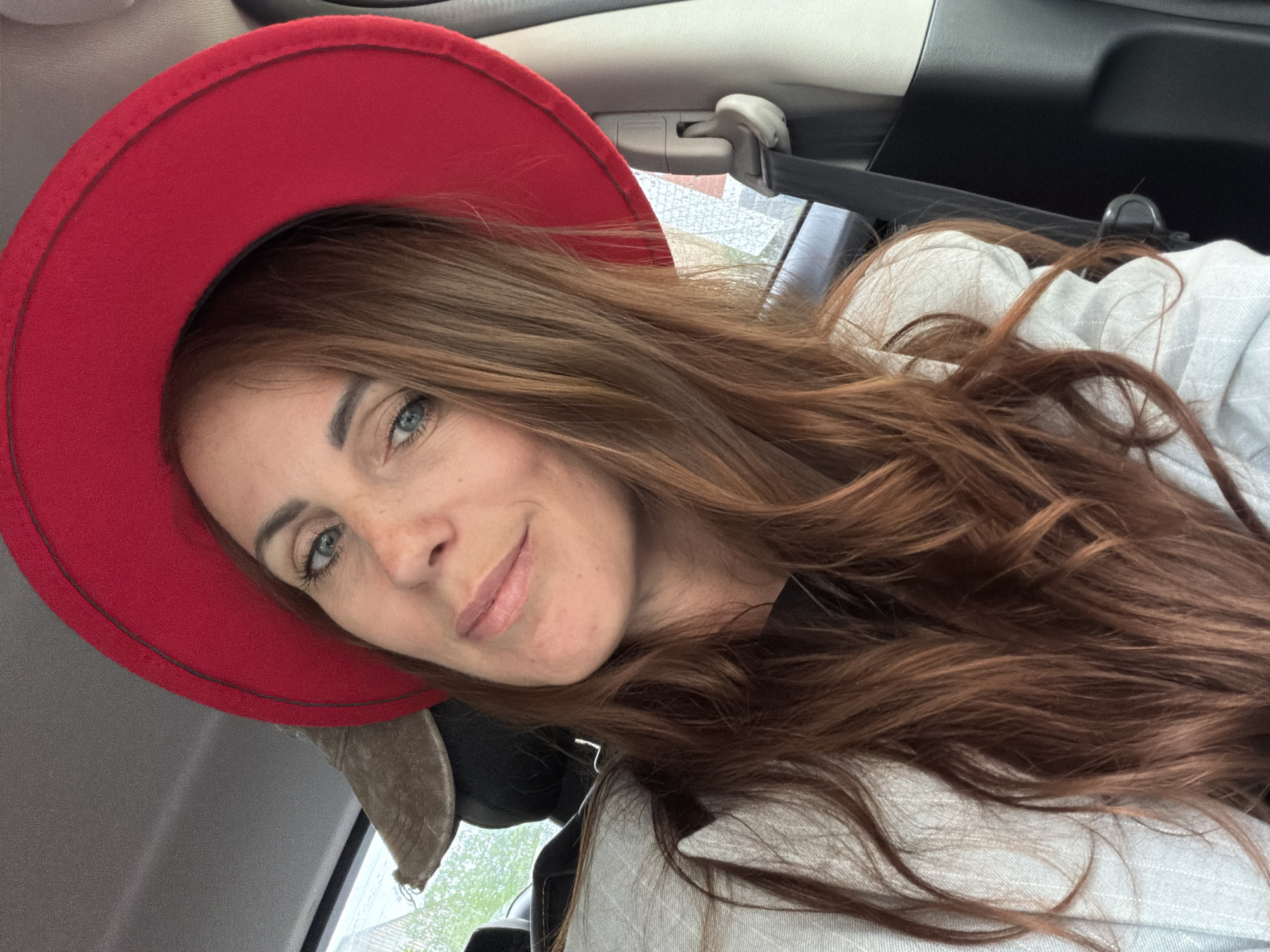 A close-up, diagonally oriented selfie features a young woman with long, wavy reddish-brown hair and bright blue eyes. She is smiling gently at the camera, her head slightly tilted. She has a fair complexion with faint freckles on her nose and cheeks. Her lips are a natural pink hue.
She wears a striking red fedora-style hat, which frames the top and left side of her face, and a light-colored, possibly white or light grey, textured shirt or jacket.
The setting appears to be inside a car, with the vehicle's headliner, various clips, and part of a window visible in the background. The outside view through the window is blurred, suggesting the car may be in motion or the focus is entirely on the subject. Natural daylight illuminates the scene, indicating it is daytime. The woman's expression conveys a sense of calm and friendliness. No visible text is present in the image. This personal portrait captures a moment within a car, situated in Birobidzhan, Russia.