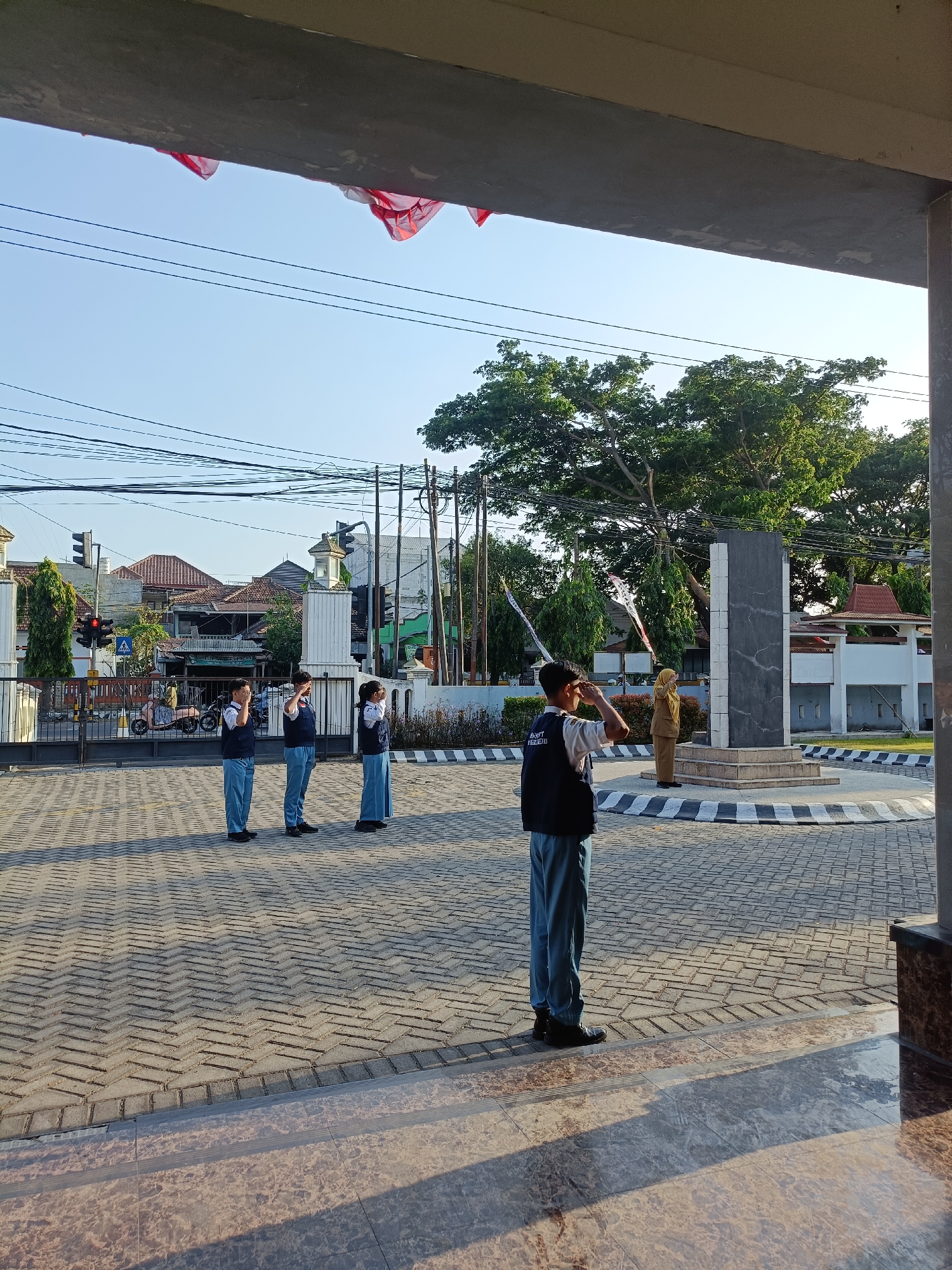 In the city of Kediri, Indonesia, at an outdoor location, several uniformed students stand at attention, saluting. The students, wearing blue vests over white shirts and light blue pants, are positioned on a paved courtyard. A woman in a tan uniform stands near a dark monument adorned with white accents. Red and white flags are visible hanging from wires. Traffic lights, buildings, and trees populate the background under a clear blue sky. The scene appears to be a formal ceremony.