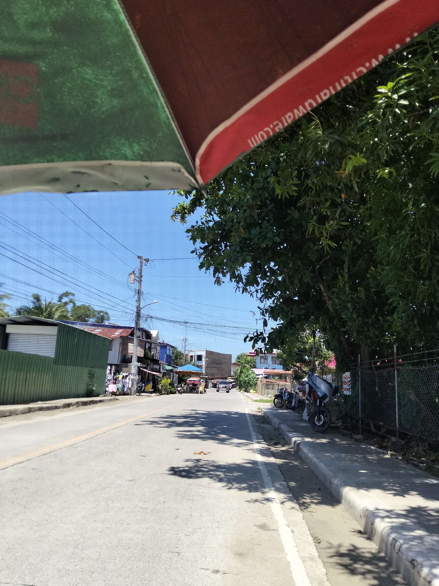 This media file depicts a street scene in Davao City, Philippines, likely during daytime on a clear, sunny day. The foreground is dominated by the underside of a vehicle, possibly a jeepney or tricycle, with a visible red and green canopy. A portion of a website address, "www.citimartware.com", is printed in white text on the red canopy.
The scene itself is a typical urban street with a paved road and a sidewalk. On the left side of the road, there is a green corrugated metal fence in front of a building with a white roller shutter door. Further down the street, more buildings are visible, including some with a rustic, corrugated metal roof and others with more modern structures. Clothes are hanging out to dry outside some of the shops, indicating a residential and commercial area.
On the right side of the road, there is a sidewalk with a shallow drainage ditch. Several motorcycles are parked along the sidewalk. A large tree with lush green foliage shades part of the sidewalk and the road. A wire fence runs along the edge of the property behind the tree.
Further down the street, traffic is visible, including cars, motorcycles, and what appears to be a yellow vehicle with a blue canopy, possibly a passenger vehicle or a food stall. Electrical wires are strung across poles, a common sight in Philippine streets. The sky is a clear, bright blue, suggesting fair weather. There are no people clearly identifiable as subjects in the image, but the presence of parked vehicles and storefronts suggests ongoing daily activity in the area. The overall impression is of a bustling, everyday scene in a Philippine city.