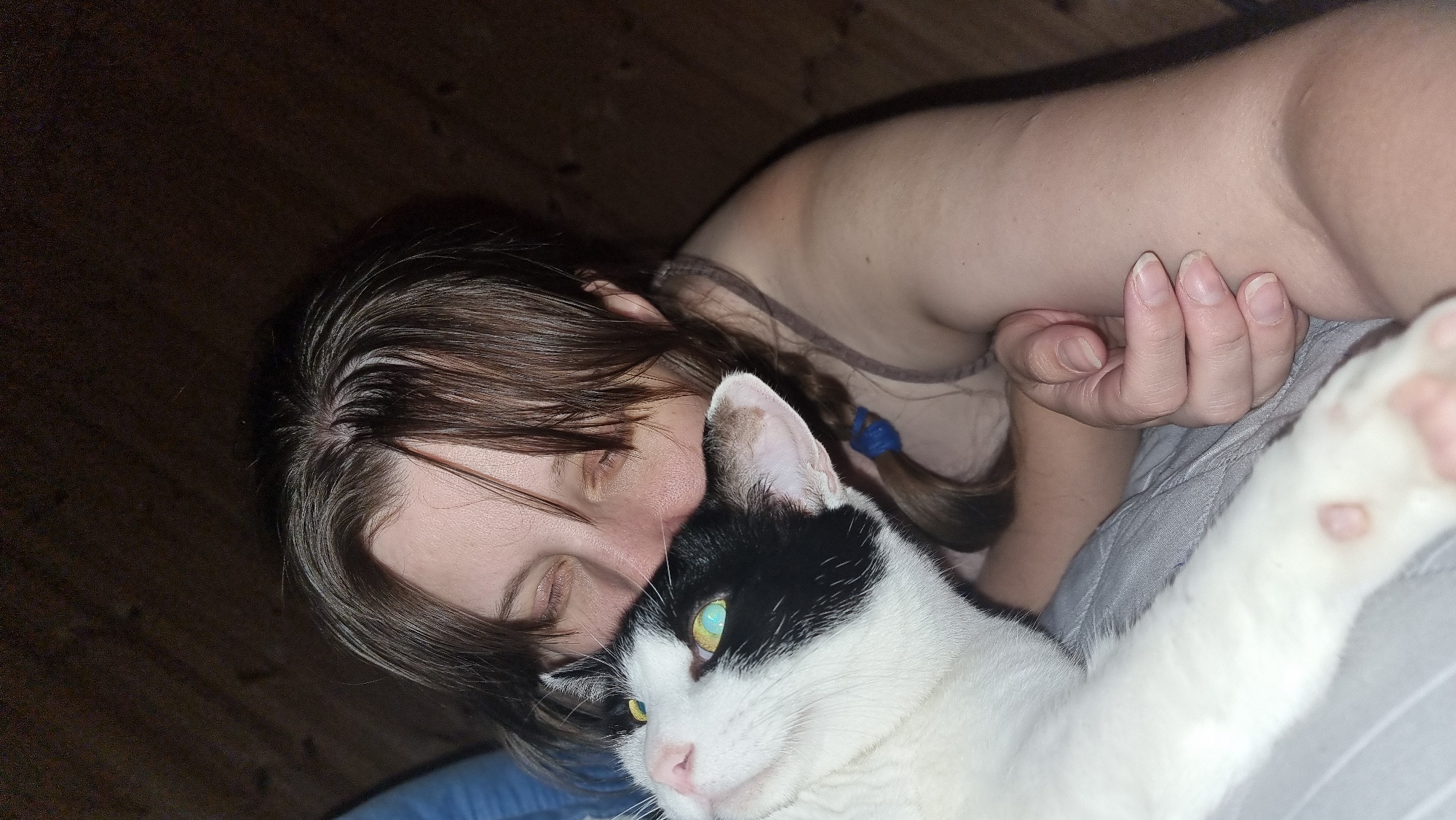 A fair-skinned person is lying on a gray bed next to a black and white cat. The person has long brown hair, part of which is braided and held with a blue hair tie. The cat's face is close to the person's, and the cat's right paw is outstretched. The location is not discernible from the image.