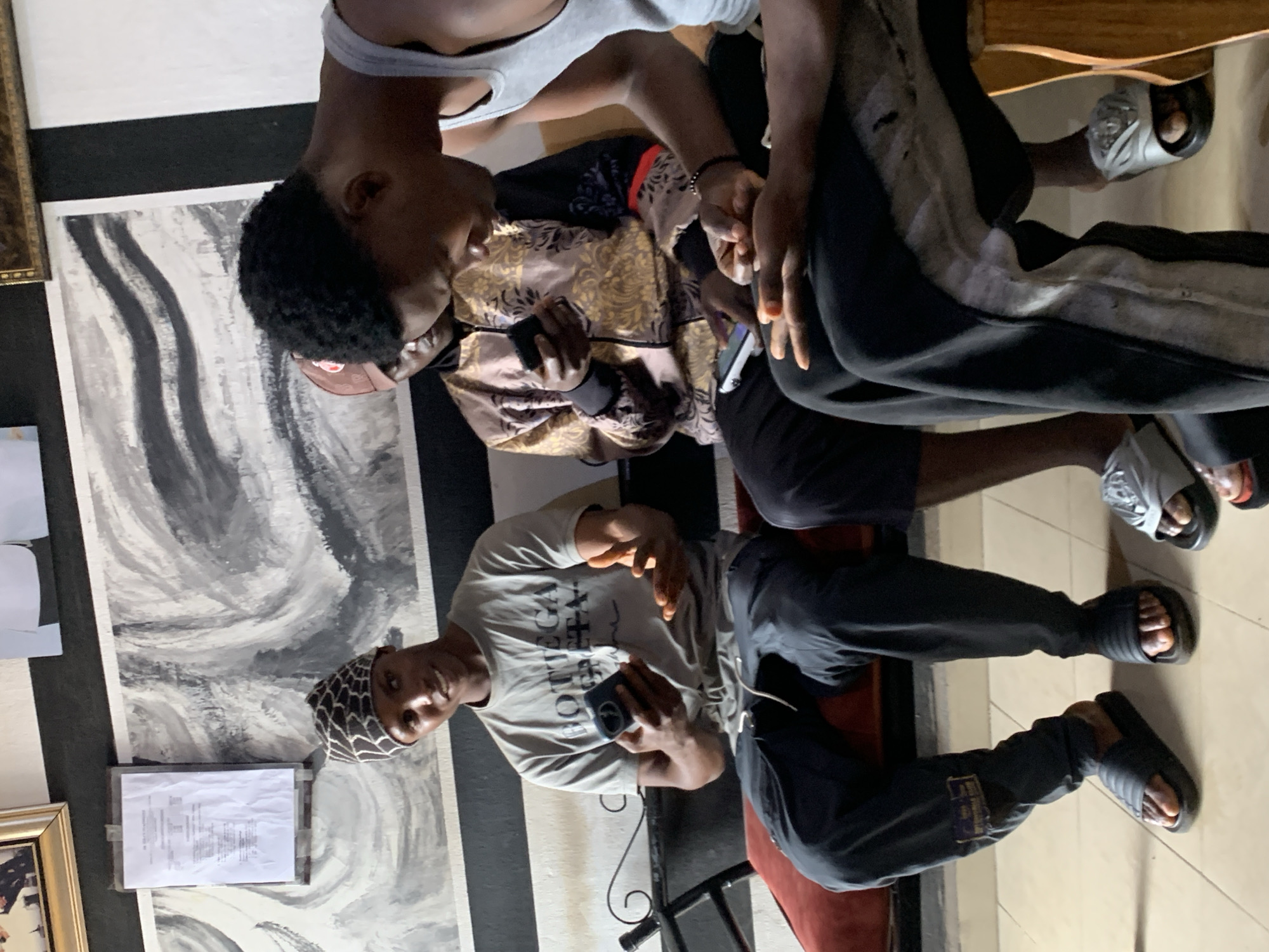 Three young men are seated on a red bench, each holding a mobile phone. The man on the left, wearing a patterned headwrap and a grey t-shirt with "BOTTEGA" printed on it, is looking at his phone and gesturing with his other hand. The man in the middle wears a brown baseball cap and a patterned jacket, and he is also looking at his phone. The man on the right, partially visible, wears a grey tank top and is looking down at his phone. They are in an indoor setting, possibly a waiting area or lounge, with a black and white striped wall behind them and an abstract painting on the upper wall. The floor is tiled. The overall scene is casual and suggests a moment of shared digital engagement. The presence of a framed document on the wall, a rolled metal object to the left, and the overall decor hint at a business or service establishment, potentially within the city of Jos, Nigeria, given the individuals' apparent ethnicity and the commonality of such establishments in urban Nigerian settings.