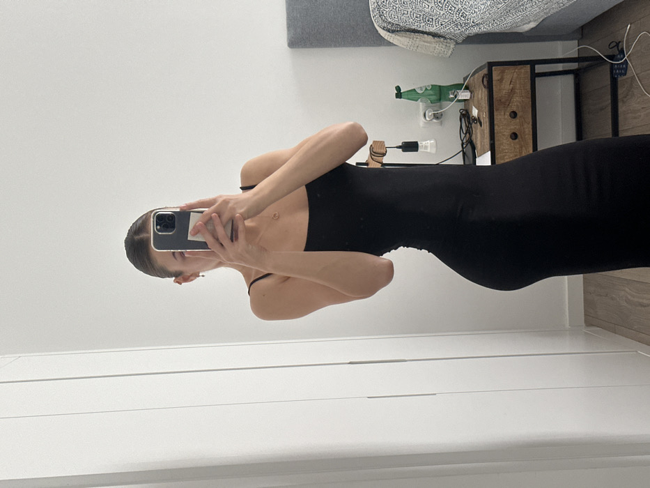 The image depicts an indoor mirror selfie, presumably taken in a bedroom located in Ericeira, Portugal. A female subject is captured from the mid-chest down, wearing a form-fitting black spaghetti-strap maxi dress. Her face is obscured by the dark-colored smartphone she holds directly in front of her to capture the reflection. She wears small earrings and a delicate necklace with a circular pendant. To the left, a white wardrobe or cabinet with vertical paneling is visible. To the right, a wooden nightstand with two dark drawer pulls and a dark metal frame stands next to a bed with a grey headboard and a patterned blanket. On the nightstand, a small wooden lamp with an exposed bulb hangs, alongside a green glass bottle, a white power outlet with a visible plug, and several small dark objects. The room features white walls and light-colored wood laminate flooring.