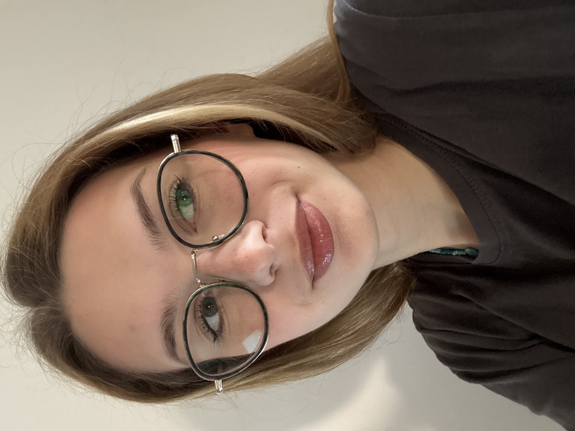 A woman with light brown hair and glasses is looking at the camera. Her eyes are of different colors, with the left eye appearing green and the right eye brown. She wears a dark-colored shirt. The background is a plain white wall. The location of the photograph cannot be determined to be Kragujevac, Serbia based on the visual information provided.