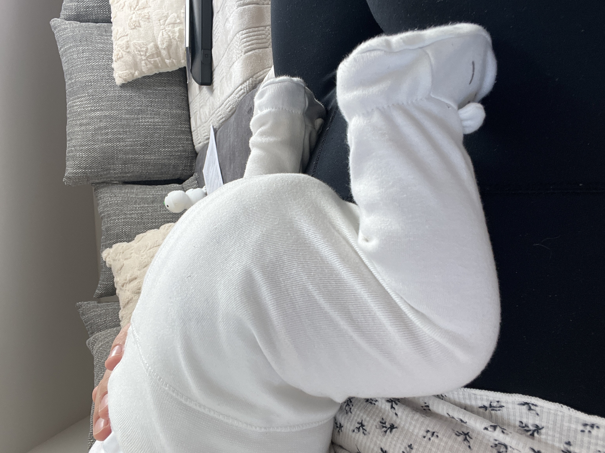 The image shows a close-up view of a baby's legs and feet dressed in white footed pajamas. The baby appears to be lying on its stomach, with one leg bent at the knee and the other extended. The background consists of a sofa with several pillows, some of which are grey and textured, and one is cream-colored with a raised pattern. A small white toy, resembling a figurine, is visible on the sofa, along with a black object that might be a tablet or laptop. There is also a sheet of paper partially visible. The overall setting suggests an indoor environment, likely a living room or bedroom, and does not provide any specific visual cues to pinpoint its location within Garmisch-Partenkirchen, Germany.