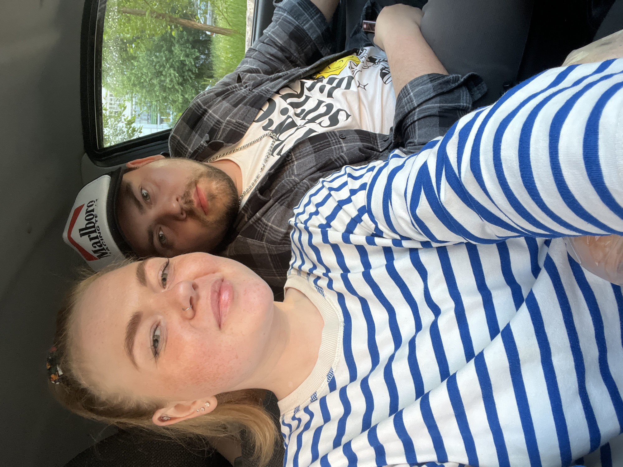 This is a casual, slightly tilted selfie of two individuals inside what appears to be a vehicle, with green foliage visible outside a window in the background, suggesting a daytime outdoor setting.
In the foreground, a female with blonde hair, a fair complexion with freckles, and light-colored eyes looks directly at the camera. She has a small ring nose piercing and multiple ear piercings. She is wearing a long-sleeved white shirt with prominent blue horizontal stripes. Her expression is relaxed and neutral.
Behind her, a male is reclining, also looking towards the camera. He has a beard and light-colored eyes. He is wearing a white t-shirt with a graphic design that includes black stylized text (partially visible, possibly "PIES?") and a yellow design. Over this, he wears an open grey and black plaid shirt. A silver chain is visible around his neck. He is also wearing a white and black baseball cap with "Marlboro" written in red, and "RACING TEAM" in smaller black letters underneath.
The scene depicts a relaxed moment, possibly during travel in Chaikovsky, Russia, as the two subjects pose for the photograph. The visible text includes "Marlboro" and "RACING TEAM" on the cap.