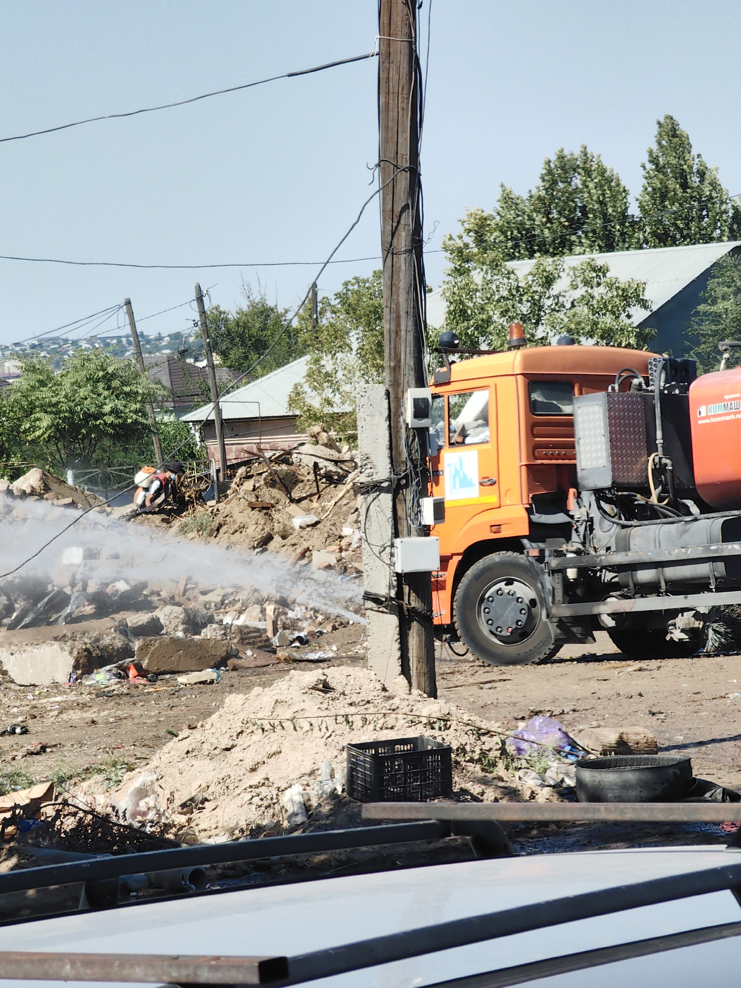 A bright orange truck with a large tank is parked on a dirt road in what appears to be a construction or demolition site. The truck is equipped with a water hose that is spraying water onto a pile of debris, which includes broken concrete and other rubble. A person in a white and orange uniform is operating the hose. In the background, there are houses, trees, and a power pole with numerous wires. The sky is clear and blue. The scene suggests ongoing work to clean up or control dust at the site. Based on the context provided by the metadata, this event took place in the city of Shymkent, Kazakhstan.