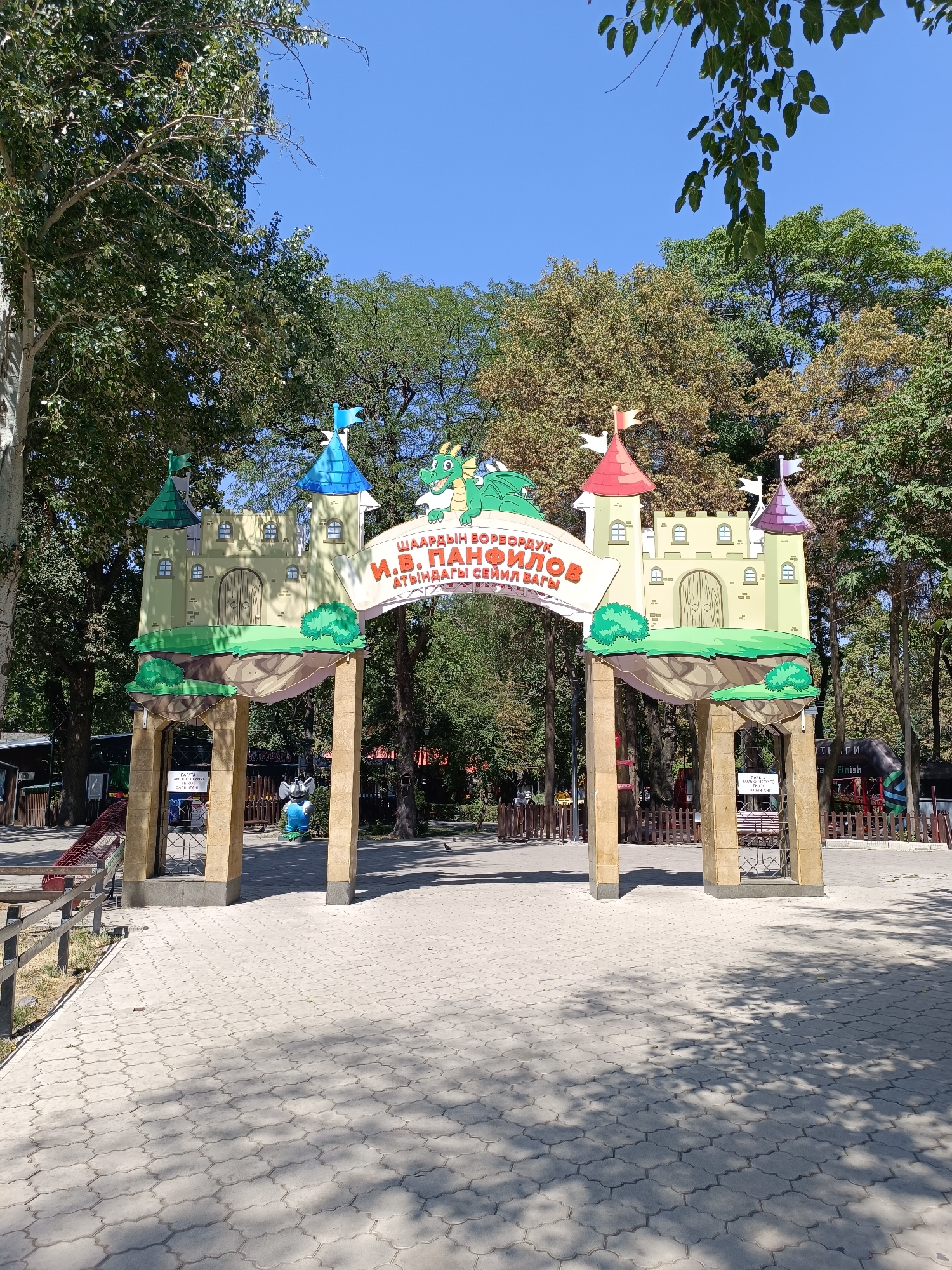 The image shows an arched entrance to a park in Bishkek City, Kyrgyzstan. The arch is decorated to resemble a castle with two towers and a dragon on top. The towers are painted in pastel colors and have flags on them. The dragon is green and appears to be breathing fire. A banner above the arch has Cyrillic writing on it. Underneath the arch, there is a paved walkway leading into the park, with trees and foliage on either side. The sky is clear and blue. The entrance appears to be part of a children's amusement park or a fairy tale-themed area.