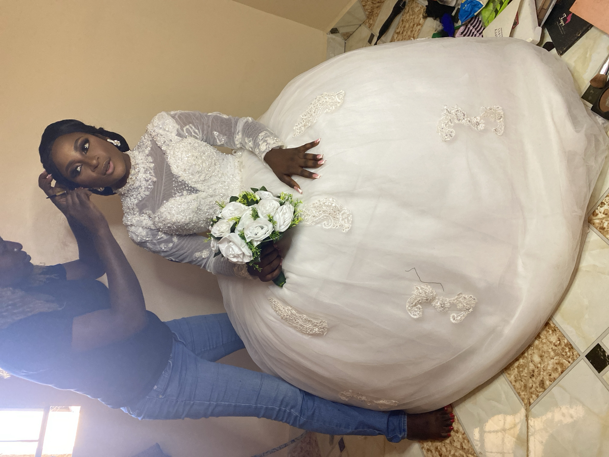 The image depicts a bride in a white, long-sleeved wedding gown adorned with lace appliques. She holds a bouquet of white roses. A person, seemingly a stylist, is attending to her hair. They appear to be indoors, possibly in a room being used for wedding preparations, with tiled flooring. Due to the angle and limited visibility, it is difficult to definitively confirm the location within Bukuru, Nigeria.