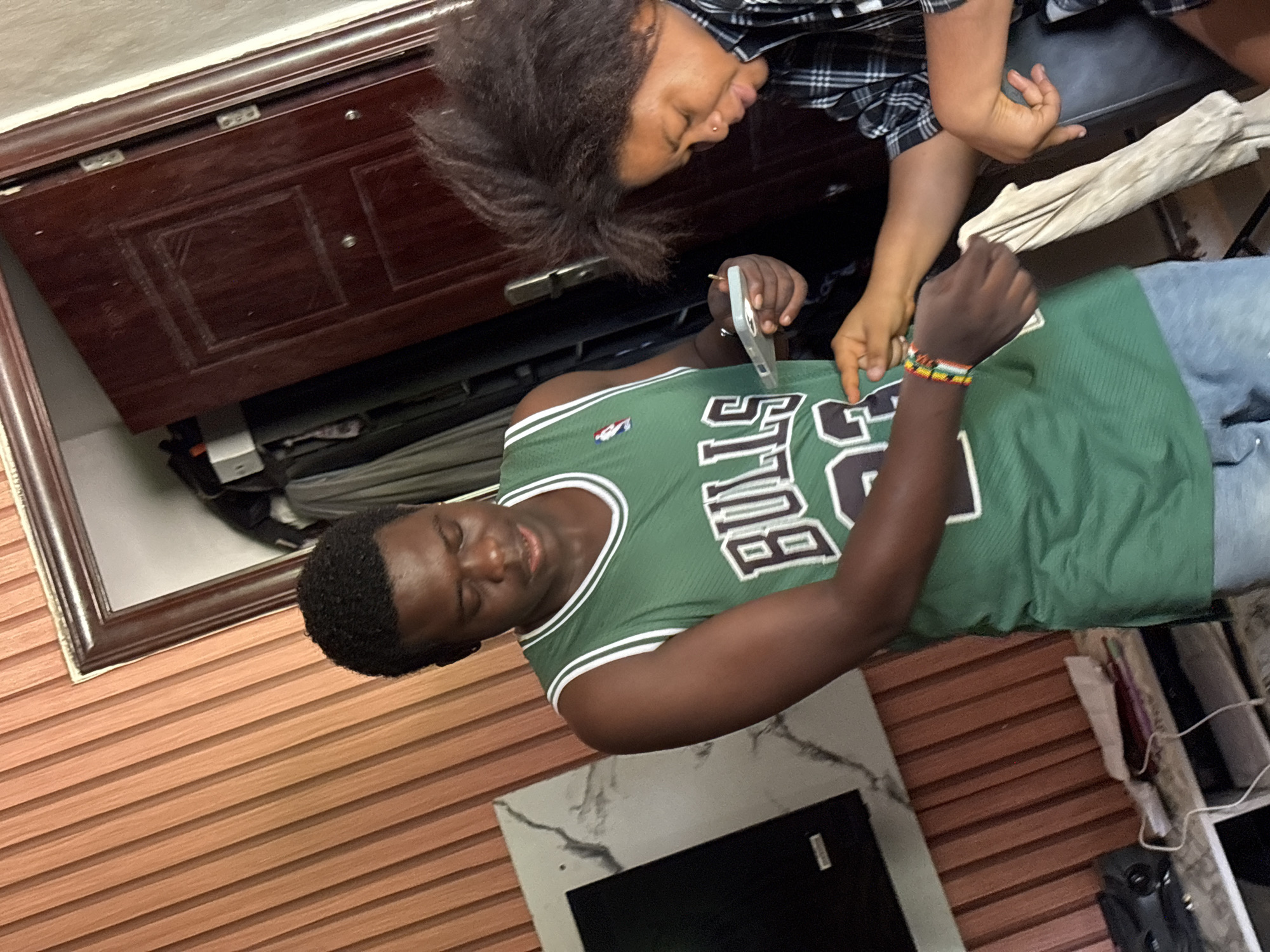 Two individuals are present, one lying down wearing a green Bulls jersey and blue jeans and the other seated and wearing a black and white plaid shirt. The seated individual is using tweezers to pluck something from the shirt of the reclining individual.
The location appears to be indoors, with a dark wooden cabinet visible in the background. A brown wooden slatted wall is also present. A small portion of Ikot Ekpene is not visible.