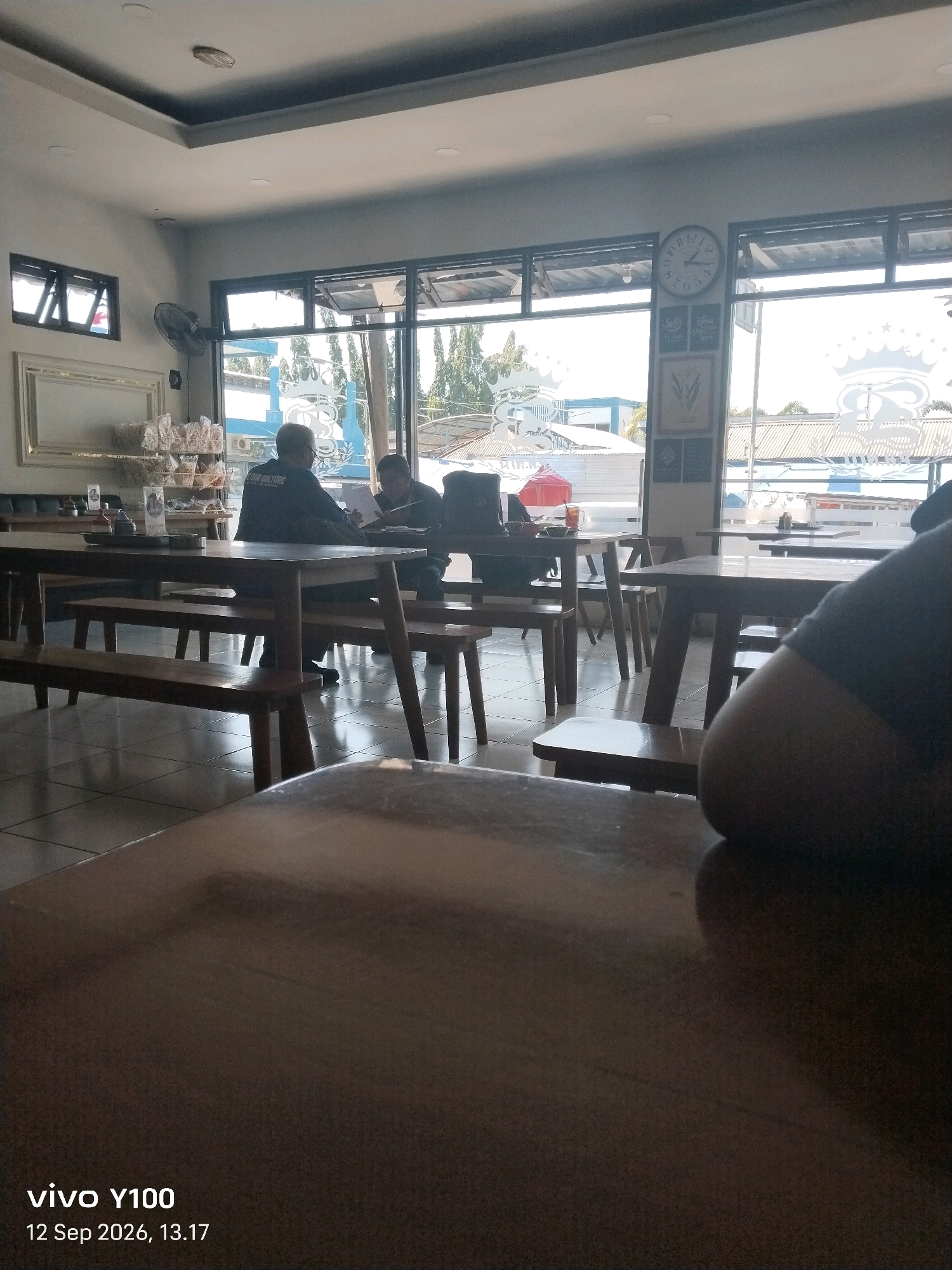 The image depicts an indoor seating area, likely a cafe or restaurant, with a bright, airy ambiance due to large windows that offer a view of the outdoors. In the foreground, a dark wooden table occupies a significant portion of the frame.
In the middle ground, two individuals are seated at a wooden table with benches. The person on the left is wearing a dark jacket with "COLE CULTURE" visible on the back. They appear to be engaged in conversation or reviewing documents with the person opposite them, who is also seated and looking downwards, possibly at a table or paper. Other tables and benches are visible, some empty, suggesting a relaxed atmosphere.
The environment includes interior elements like a ceiling with recessed lighting, a wall-mounted fan, and a large framed decorative piece. Through the large windows, an outdoor scene is visible, featuring trees and buildings, with a prominent clock on the interior wall. The overall setting suggests a public establishment in Situbondo, Indonesia, possibly a roadside eatery or cafe given the architectural style and the glimpse of the outdoor environment. The presence of packaged snacks on shelves in the background further supports this interpretation. The image is dated "12 Sep 2026, 13.17" and branded "vivo Y100".