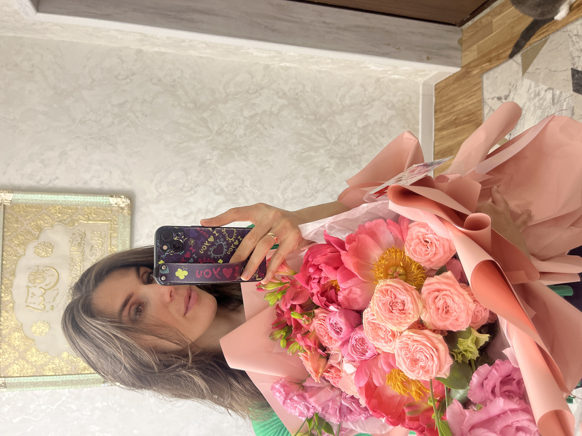 A woman is taking a selfie with a bouquet of flowers. She is holding her phone in her right hand, and the bouquet is in her left arm. The flowers are a mix of pinks, oranges, and yellows and are wrapped in pink paper. The woman has brown hair and is wearing a green top. The background shows a textured wall and a mirror. Location is the city of Kaspiysk, Russia.