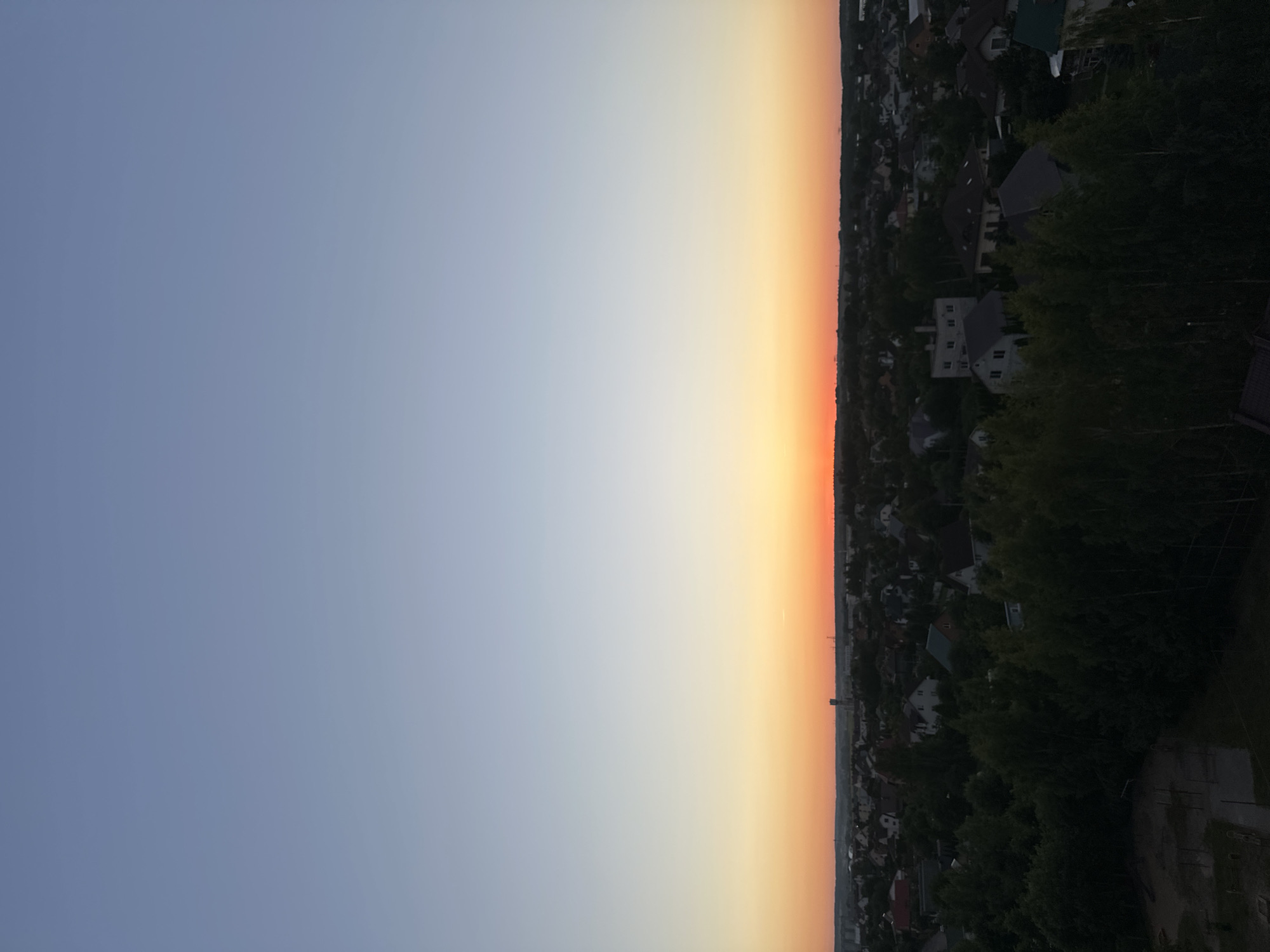 The image captures a wide, panoramic view of a suburban landscape at dusk, likely within or near Шчомысліцкі сельскі Савет, Belarus. The upper two-thirds of the frame is dominated by a clear sky, transitioning from a pale blue at the top to a soft gradient of yellow and orange hues near the horizon, indicating the setting sun.
The lower third of the image reveals a dense residential area characterized by numerous houses, varying in architectural style and size. Many of these houses are surrounded by mature trees, creating a lush, green canopy that partially obscures the rooftops. Some of the trees are quite tall, their branches extending upwards towards the sky.
In the foreground, on the left side, a small playground or sports area is visible. It appears to be an outdoor facility with some rudimentary structures, possibly swings or exercise equipment, situated on a sandy or gravel surface. The lighting in this area is dim, suggesting it is shadowed by larger trees or buildings.
Further into the residential area, several houses are more clearly discernible. These range from what appear to be single-family homes with pitched roofs to larger, multi-story buildings. The overall impression is of a well-established suburban community nestled amongst abundant greenery.
In the distance, along the horizon, faint structures of utility towers or industrial elements can be spotted, breaking the uniformity of the landscape. The scene is peaceful and serene, characteristic of a quiet evening in a rural or suburban setting. No people are visible in the frame.