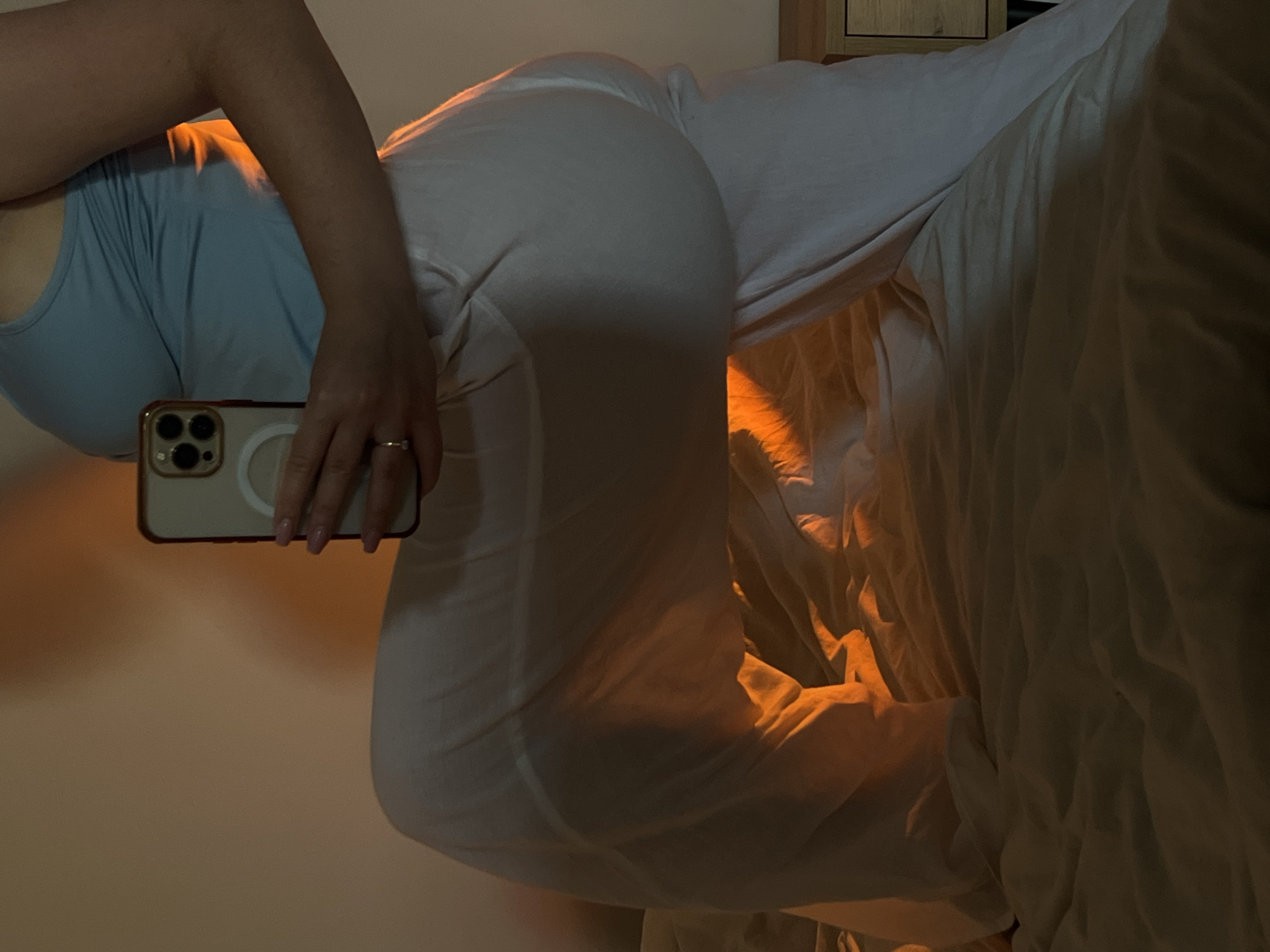 The image shows a person partially visible from the torso up, wearing a light blue tank top and white pants. The person is holding a smartphone with a triple-lens camera, presumably taking the photo. The background features a bed with crumpled white sheets, creating a sense of domesticity. A cabinet can be seen in the upper right corner. The lighting appears warm, with orange hues, possibly due to an indoor light source or sunset. There is no information to indicate the location in the city of Ternopil, Ukraine.