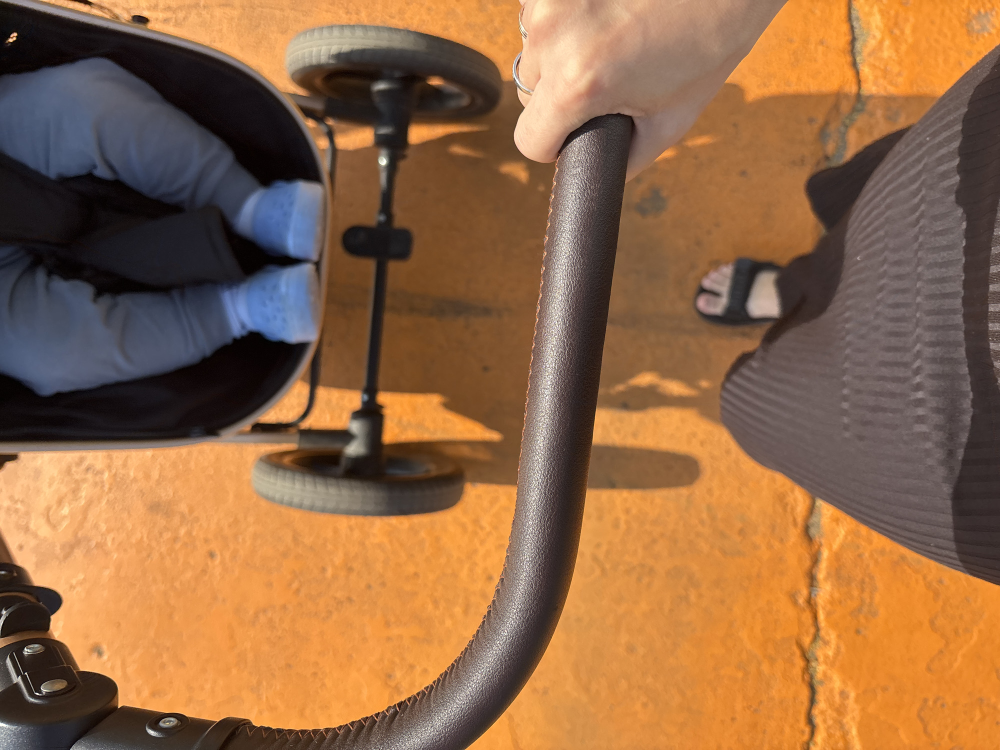 The image shows a first-person perspective of someone pushing a stroller on an orange, textured surface, possibly asphalt or concrete. The stroller contains the legs of a young child wearing light blue shoes and gray pants. The person pushing the stroller is wearing a dark brown, ribbed skirt and black sandals. Their right hand, adorned with two silver rings, is gripping the stroller's handle. The scene appears to be outdoors in daylight, casting shadows from the stroller and the person. The overall environment suggests a pedestrian area within the city of Naberezhnye Chelny, Russia.