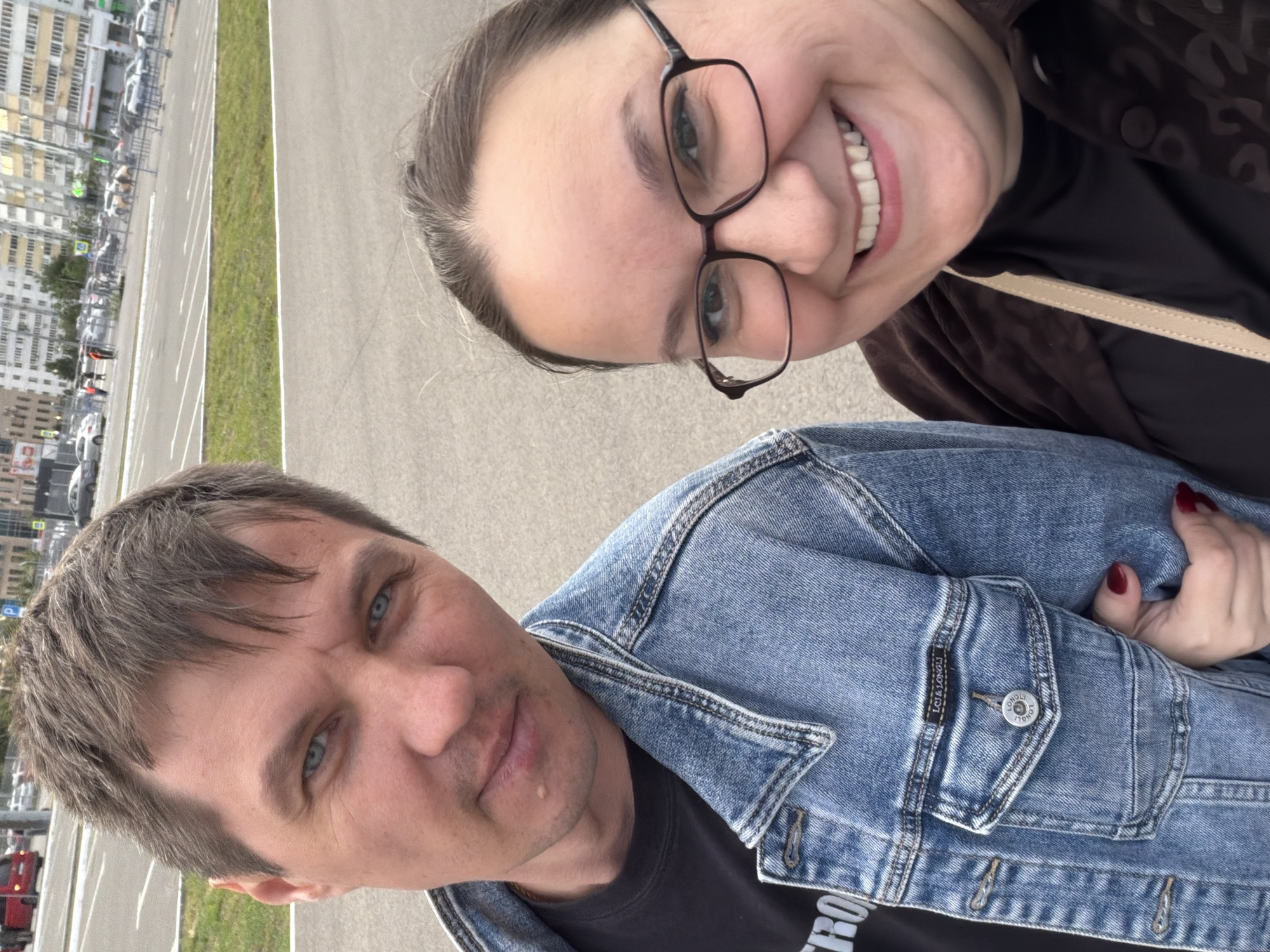 The image captures two individuals in a selfie. A woman with glasses smiles broadly, while a man in a denim jacket stands slightly behind her. In the background, a parking lot and buildings are visible, suggesting an urban environment. Based on available data, this scene is located in the city of Kazan, Russia.
