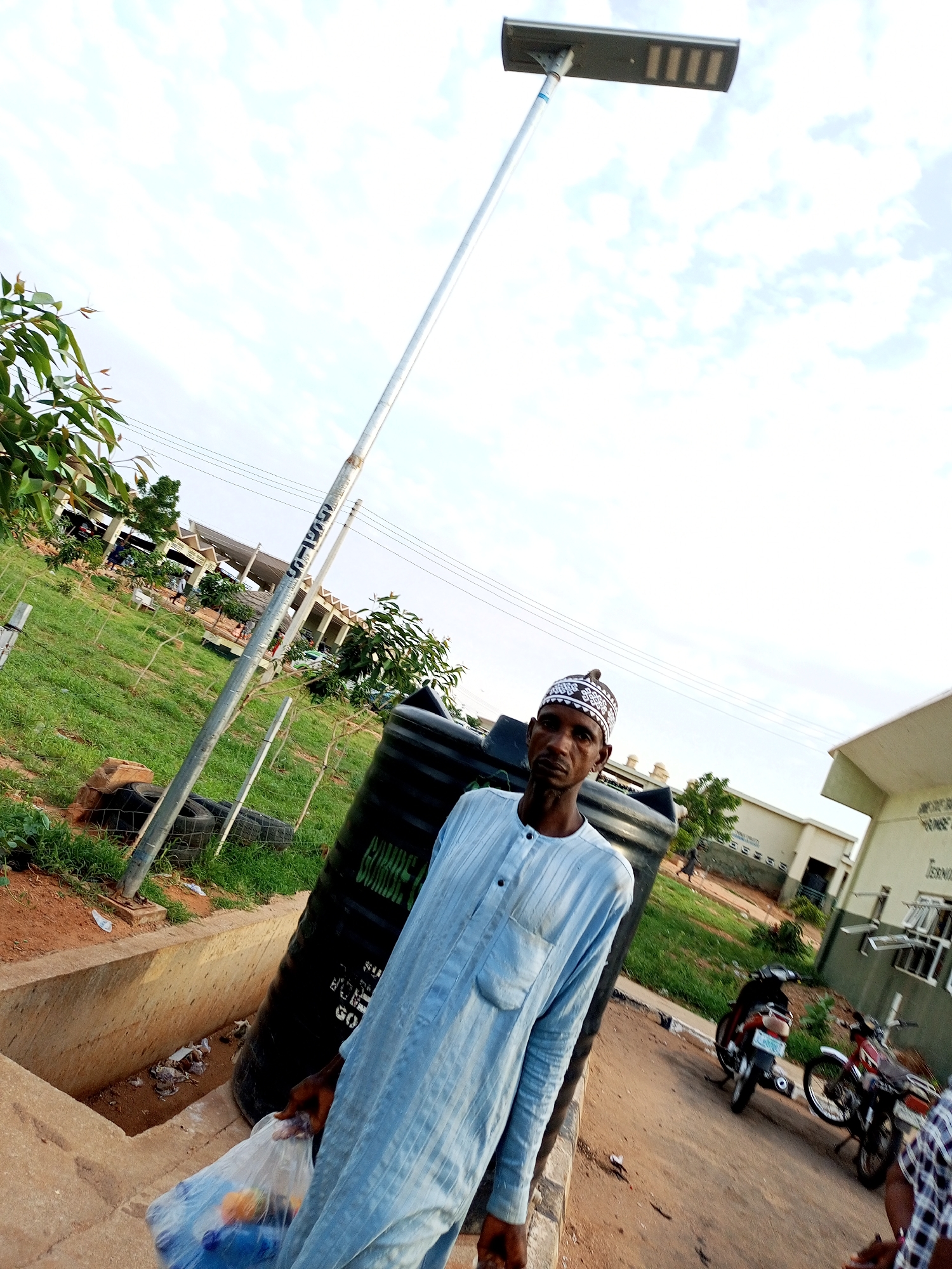 In Gombe, Nigeria, a man wearing a light blue traditional outfit and a white and black patterned cap stands near a tall solar-powered streetlight. He is holding a clear plastic bag filled with items. To his right, a large black water tank is visible. In the background, there are several buildings with green trim, grassy areas, and a dirt road with two motorcycles parked on it. The sky is partly cloudy.