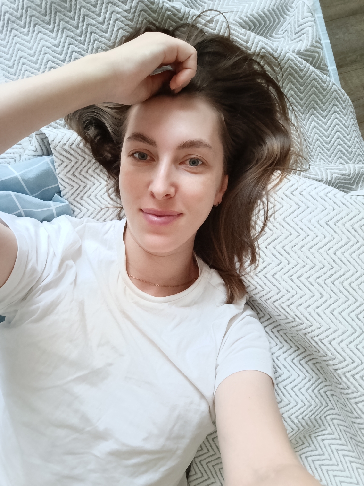 A woman is lying on a bed, wearing a white T-shirt. She is looking directly at the camera with a slight smile. Her hair is brown and disheveled, with one hand resting on her forehead. The bed has a grey and white patterned cover. A blue patterned pillow is visible on the left side. The location is the city of Chaikovsky, Russia.