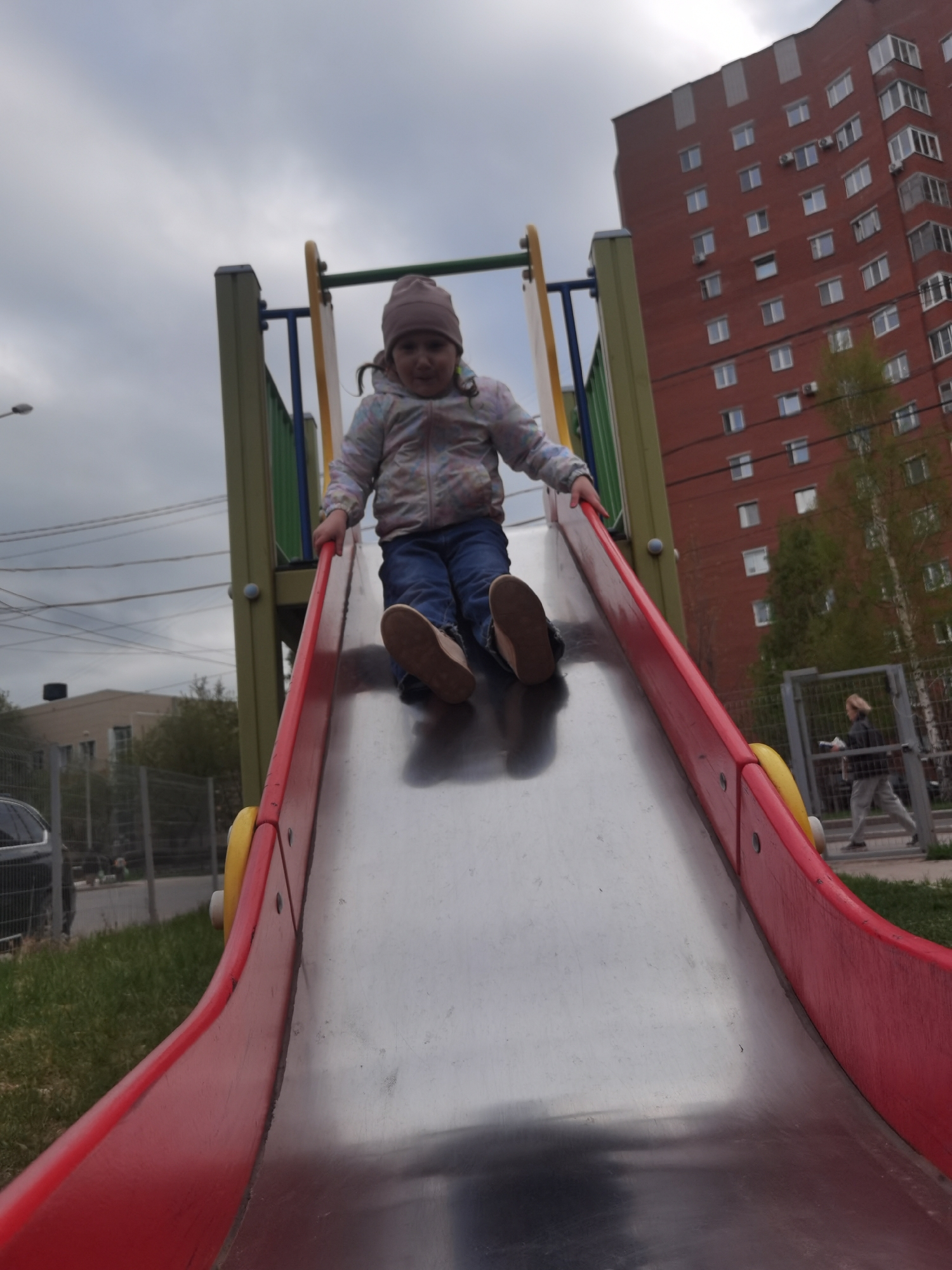 A young girl is sliding down a red and silver slide in Perm, Russia. She wears a light jacket, blue jeans, brown shoes, and a brown beanie. The slide is part of a playground structure with green, blue, and yellow accents. Behind the playground is a large red brick building. A person walks by on a path near a gray fence. The sky is overcast. There is grass around the playground equipment. A car is parked in the distance.