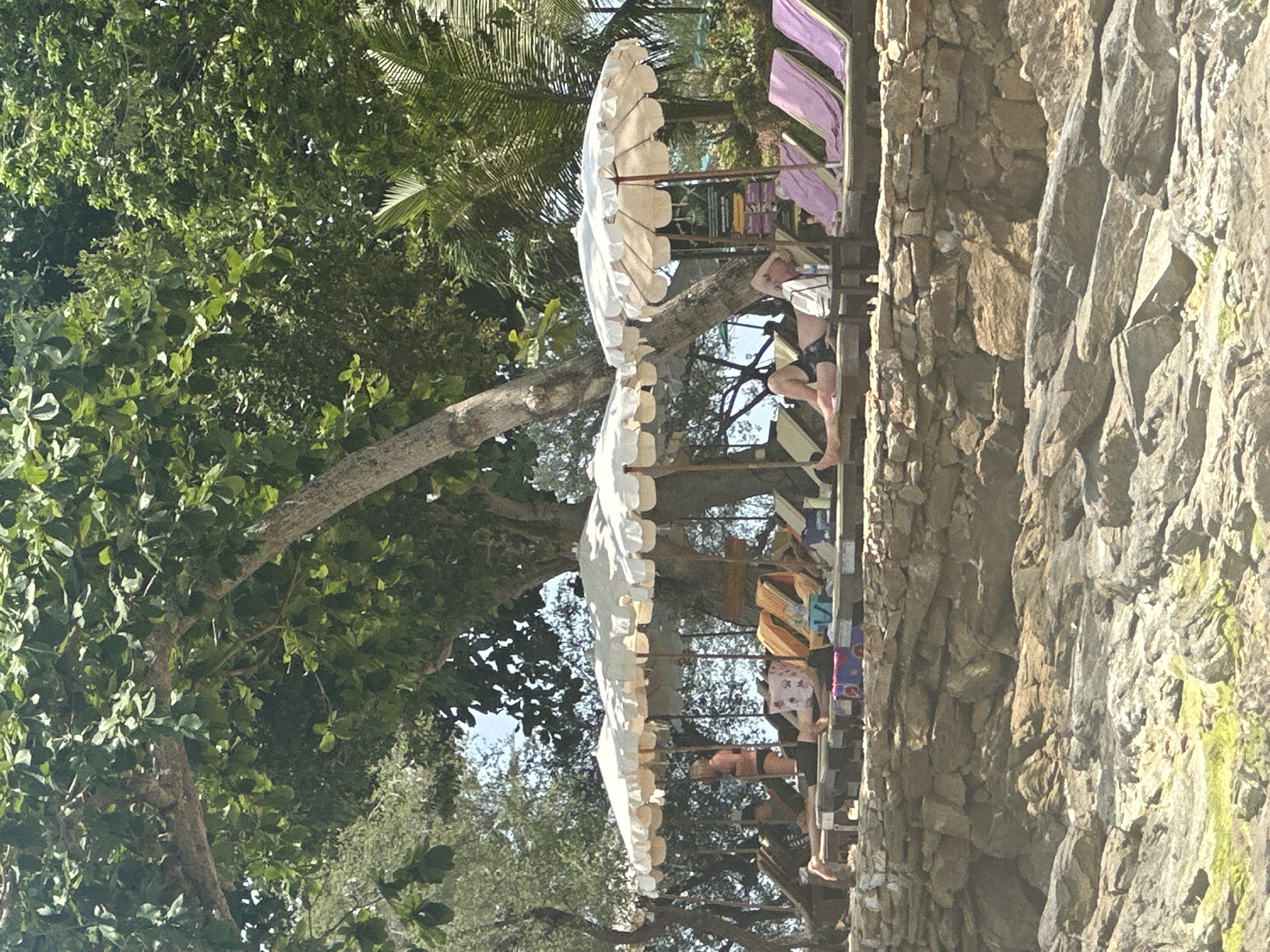 The image shows a tiered seating area on a rocky cliff face in Ao Hin Khok, Thailand. The seating area has multiple levels with lounge chairs shaded by large, white umbrellas with scalloped edges. People are relaxing in the chairs. The left side of the frame is filled with lush green trees and foliage, while the right side is dominated by the rocky cliff.