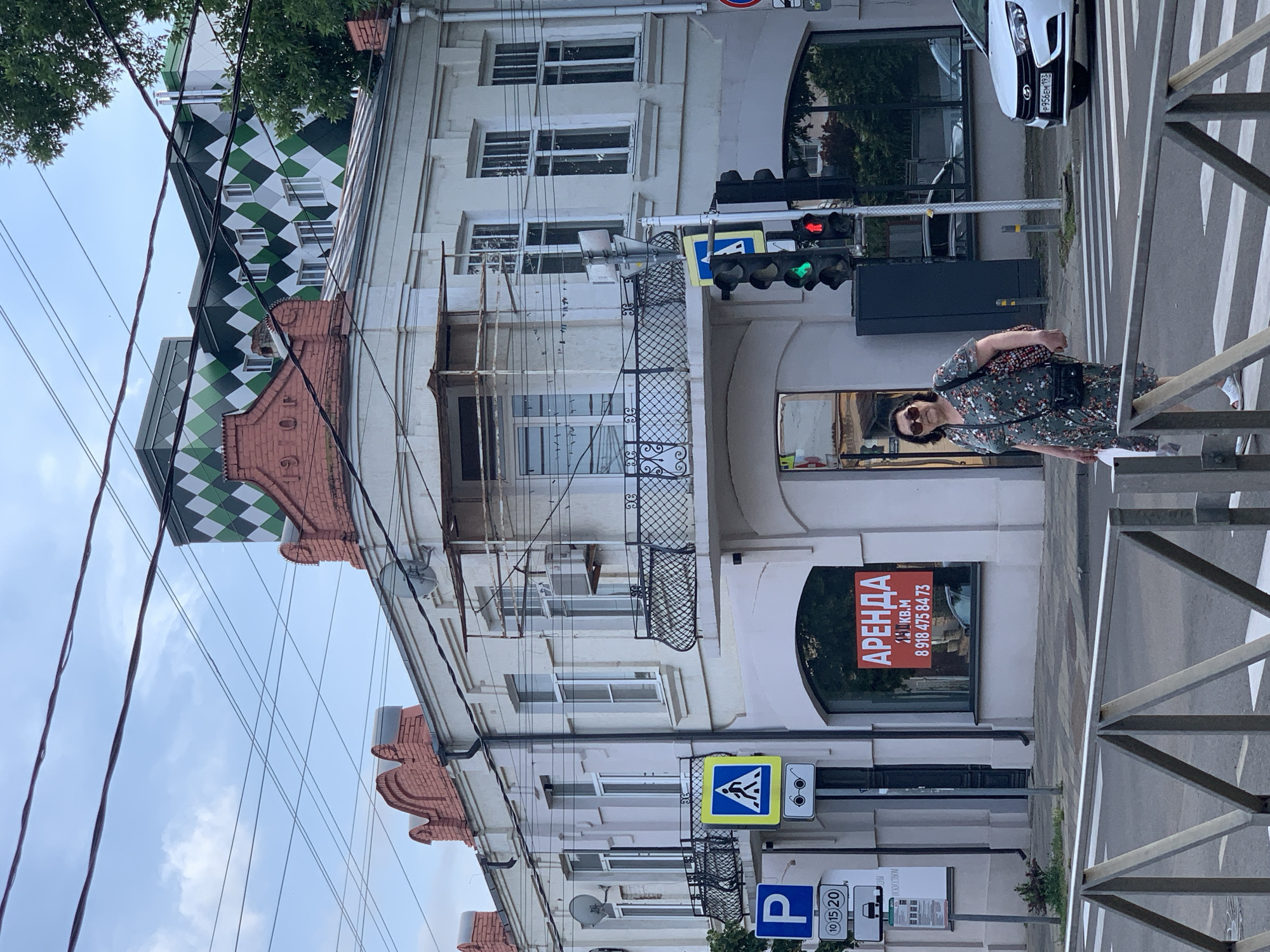 A woman in a floral dress walks across a pedestrian crossing. She wears sunglasses and carries a purse over her shoulder and a white bag in her right hand. She is walking in front of a light-colored building with a red sign that says "АРЕНДА" (rent) in Russian. A traffic light is visible with a green light illuminated. In the background, a white car with a license plate ending in "193" drives past. The scene is set in an urban environment with overhead power lines crisscrossing the sky.