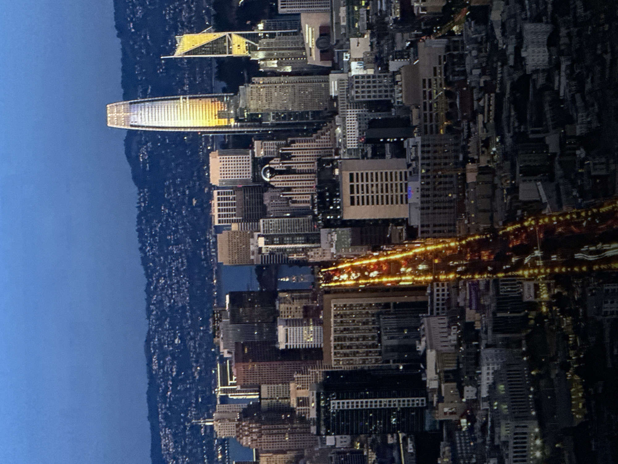 This is an aerial, nighttime photograph of a cityscape. The sky is a deep blue, transitioning to a lighter blue near the horizon where a range of hills is visible. The city is densely populated with numerous buildings of varying heights, many of which are illuminated. A prominent feature of the cityscape is a series of tall skyscrapers, including one exceptionally tall, slender building with a shimmering facade that reflects golden light. To the right of this tall building is another distinctive skyscraper with a triangular, golden-hued top. A wide, illuminated road with streaks of light, suggesting moving vehicles, cuts diagonally through the lower portion of the image, creating a vibrant contrast with the darker city structures. The overall impression is one of a bustling metropolis at dusk or early night. The scene is not located in Saratov, Russia.