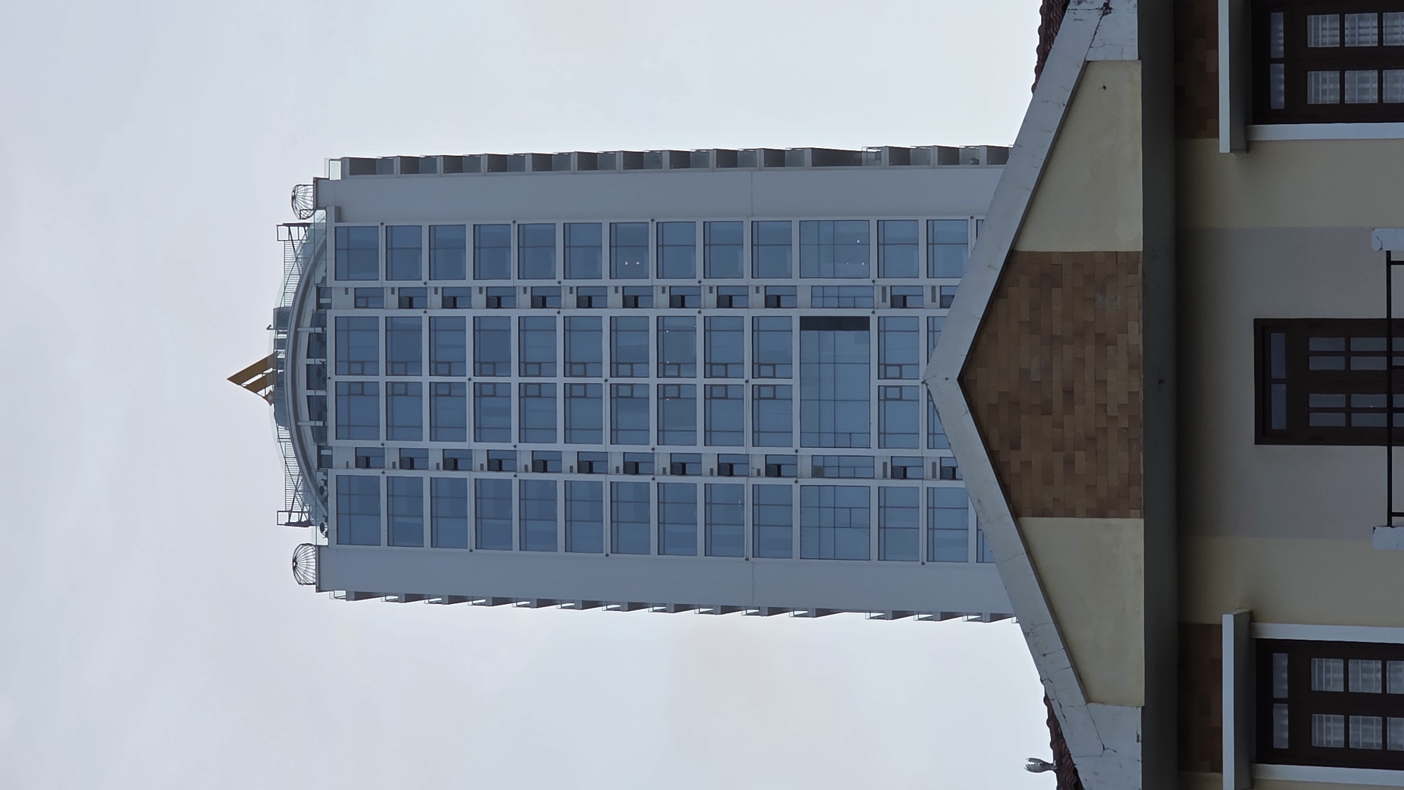 The image captures a vertical view of architectural elements in Nha Trang, Vietnam. A modern high-rise building with numerous windows is juxtaposed with a section of an older, low-rise structure. The high-rise is a light color, featuring a rounded top adorned with a gold-colored decorative element, possibly a logo. The windows appear reflective, suggesting they are made of glass. To the right of the high-rise is a portion of a building with visible windows and a peak design made of brick and stucco, indicating a different architectural style and age. The sky is overcast and provides the backdrop for the scene.