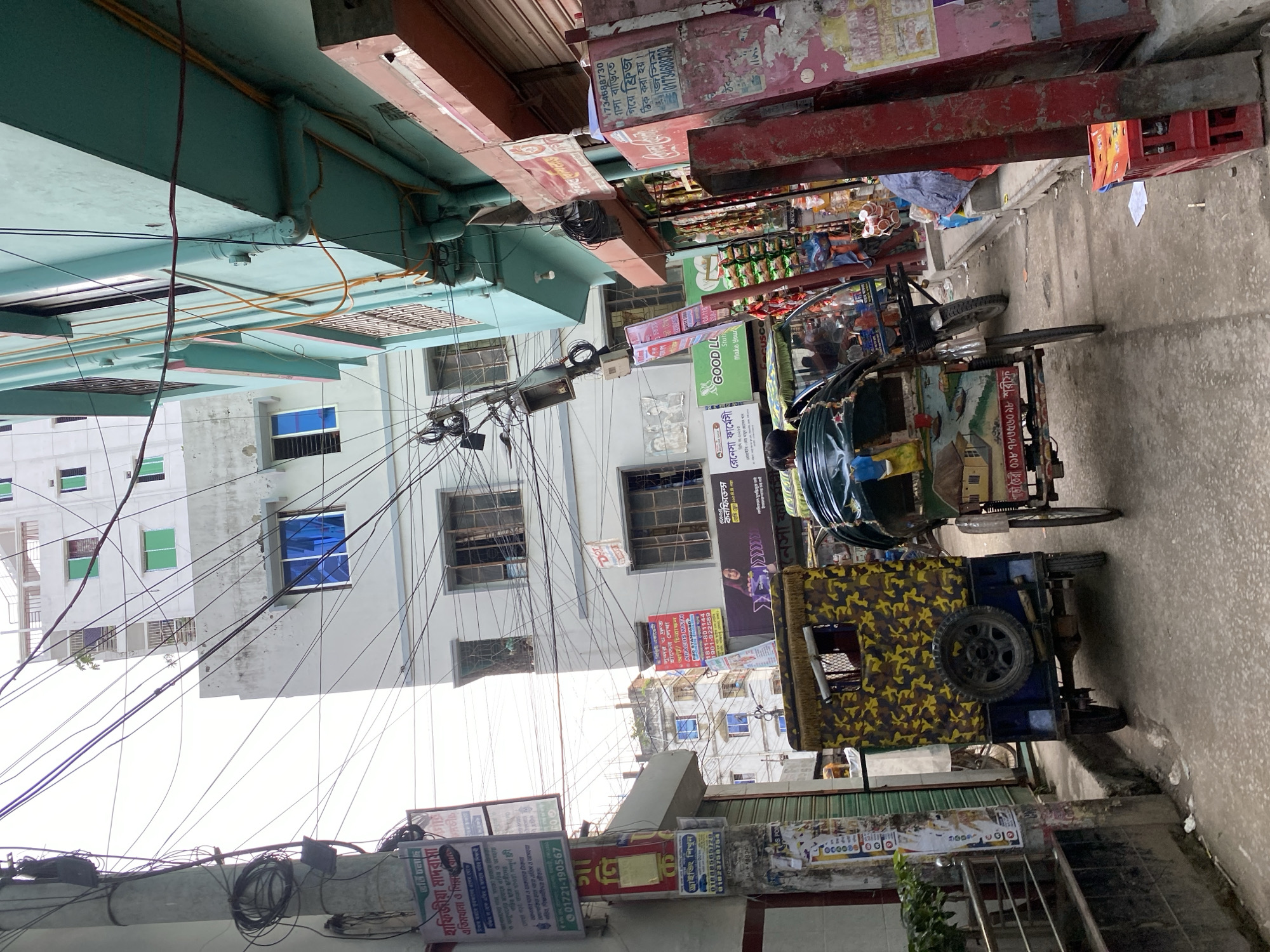 The image depicts a busy street scene in Dhaka, Bangladesh, likely in a commercial or residential area given the mix of buildings and pedestrian activity. In the foreground, two auto-rickshaws, known locally as CNGs, are parked on the street. The first auto-rickshaw, on the left, is painted with a camouflage pattern of yellow and green, with a spare tire mounted on its rear. The second auto-rickshaw, on the right, is a more traditional model, also carrying passengers and goods, with a driver visible.
The street is lined with multi-story buildings, some appearing to be apartments or commercial establishments. The architecture is a mix of concrete structures, with many windows and balconies. Several signs in Bengali are visible on the buildings and lampposts, indicating businesses, educational institutions, and advertisements. The buildings have a somewhat worn appearance, with peeling paint and visible signs of wear and tear, common in urban environments.
Overhead, a complex network of electrical wires crisscrosses the sky, a characteristic feature of many streets in Dhaka. This creates a dense and somewhat cluttered visual aesthetic. The ground is a paved street and sidewalk, with some debris and puddles suggesting recent rain or general urban conditions.
In the background, the sky is overcast, contributing to the diffused lighting of the scene. The overall atmosphere is one of a bustling, everyday urban environment, characteristic of Dhaka's vibrant street life. The image captures a slice of life in the city, showcasing its modes of transport, architecture, and the dynamic, energetic ambiance.