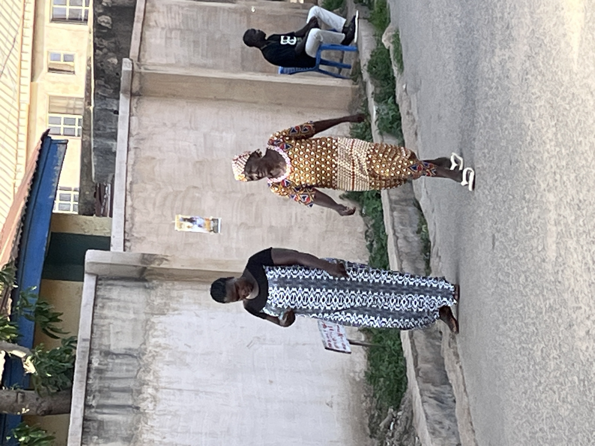 A daytime, eye-level shot depicts a street scene in Jos, Nigeria. Two women walk in the street, while a man sits on a blue chair against a building. The woman closest to the camera wears a long, black and white patterned dress. The other woman wears a brown and tan patterned dress and white slippers. The man is wearing a black t-shirt, grey pants, and black sneakers. Behind the pedestrians is a concrete wall with vegetation at its base. Buildings are seen to the left. The road surface is visible and dry.