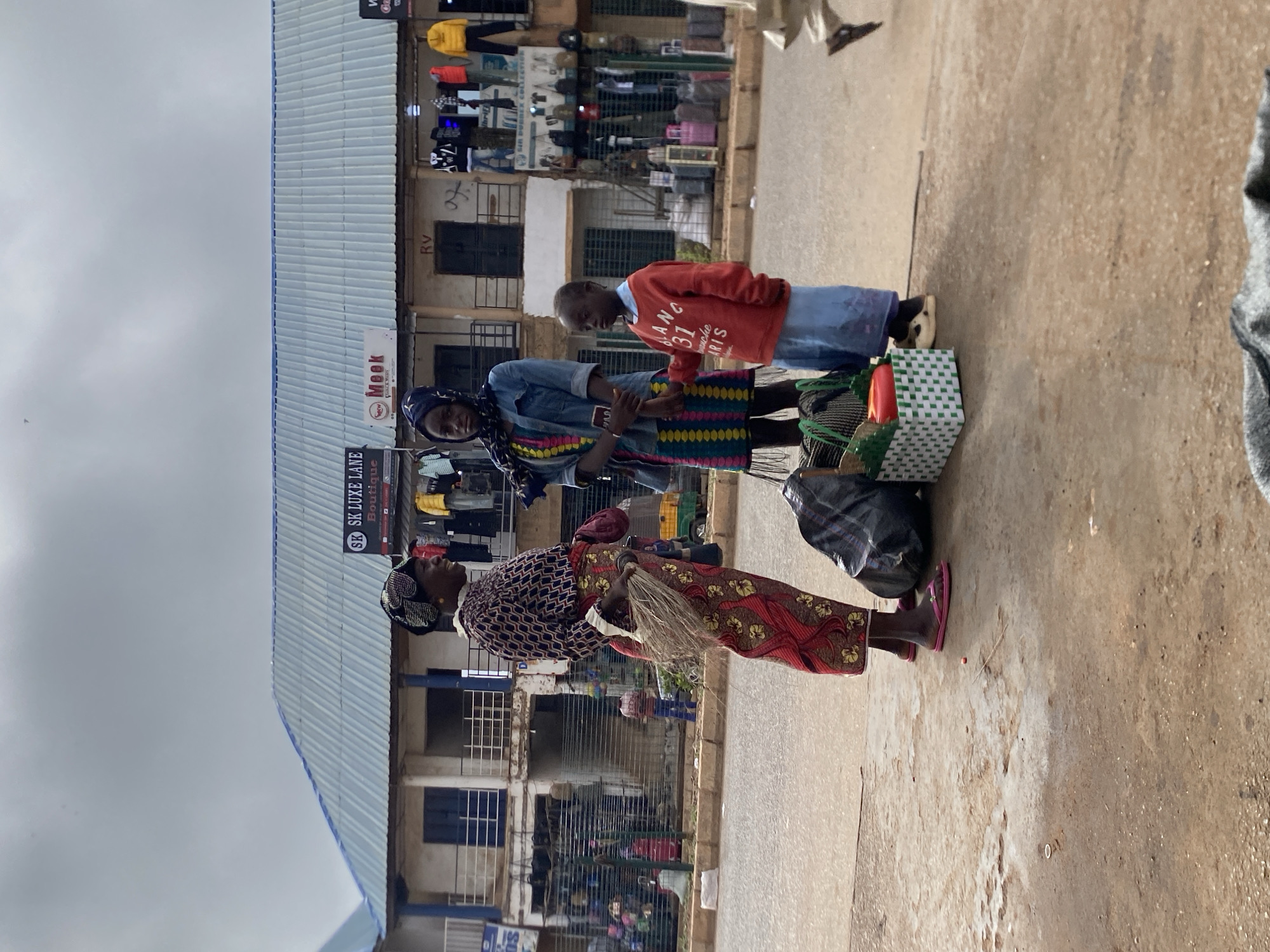 Here is a concise, forensic description of the scene:
**Location:** Bukuru, Nigeria
**Subjects:**
* Three individuals are standing on a street.
* An older woman is wearing a patterned dress, head covering, and flip-flops. She is carrying a straw object.
* A second woman wears a denim shirt, head covering, and a patterned skirt. She holds what appears to be a phone or electronic device in her hand.
* A child in a red sweater and blue pants stands close to the second woman.
* Several bags or bundles rest on the ground near them.
**Objects:**
* Bags of various materials and patterns.
* A broom or whisk made of natural fibers held by the older woman.
* Buildings with multiple levels. A sign for "SK LUXE LANE Boutique" is visible. Clothing is displayed in a shop front on the upper level.
**Actions:**
* The two women are likely engaged in a transaction or discussion. The child seems to be observing.
**Interactions:**
* The three people seem to form a familial or economic unit, possibly shopping or conducting business.
**Environment:**
* The scene is an outdoor street with buildings and shops in the background. The ground appears to be a paved surface. The sky is overcast. The location is a commercial zone with shops and potentially vendors.